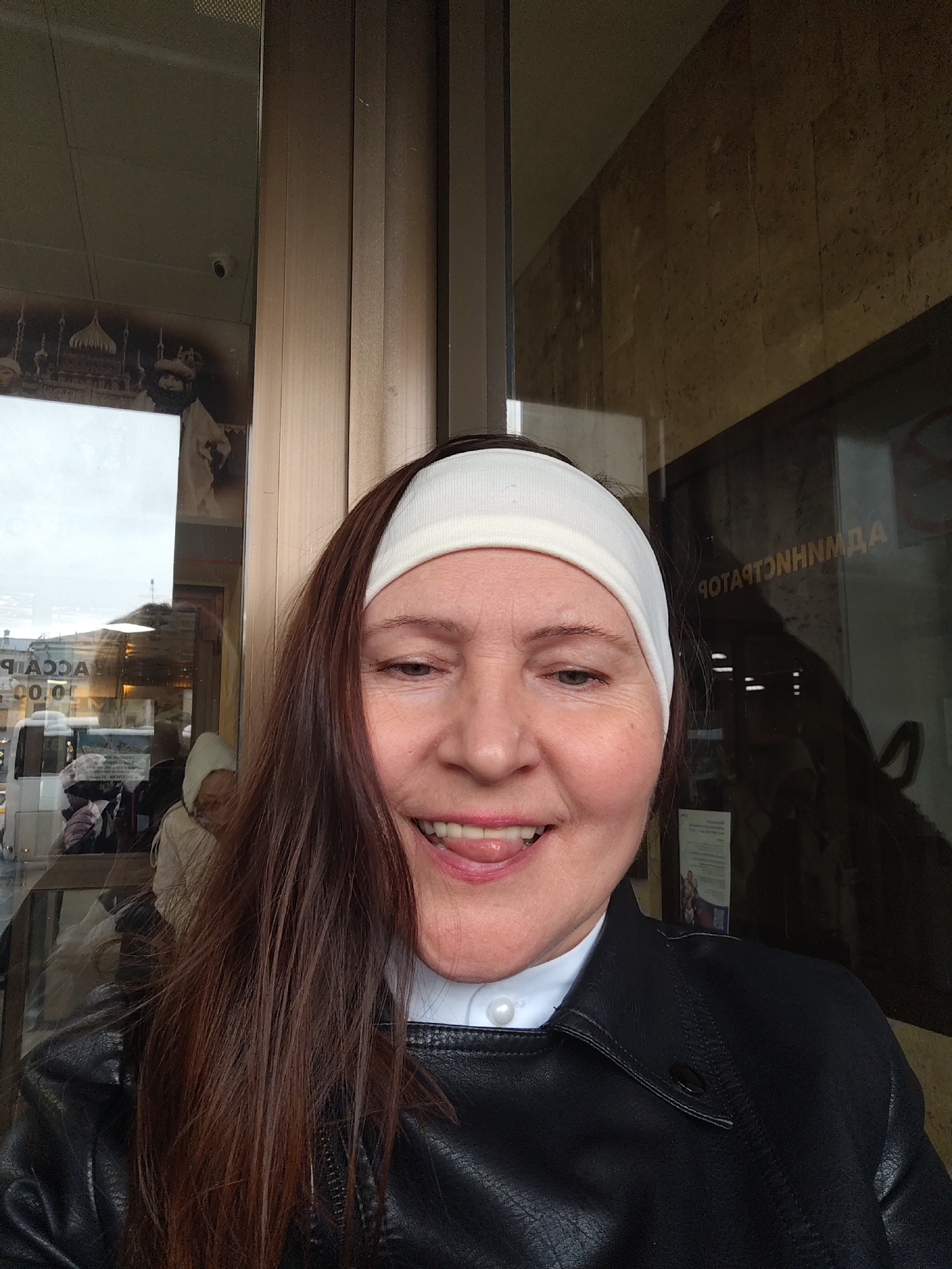 A woman is standing in front of a glass window. She has a white headband on her head and long, dark hair. She is wearing a black leather jacket over a white collared shirt. The woman is sticking her tongue out and smiling. In the background, there is a reflection of a street scene with people and buildings. The words "ACCA" are visible in the reflection. The sign behind the glass says "АДМИНИСТРАТОР". Based on the reflection, this is in Moscow, Russia.