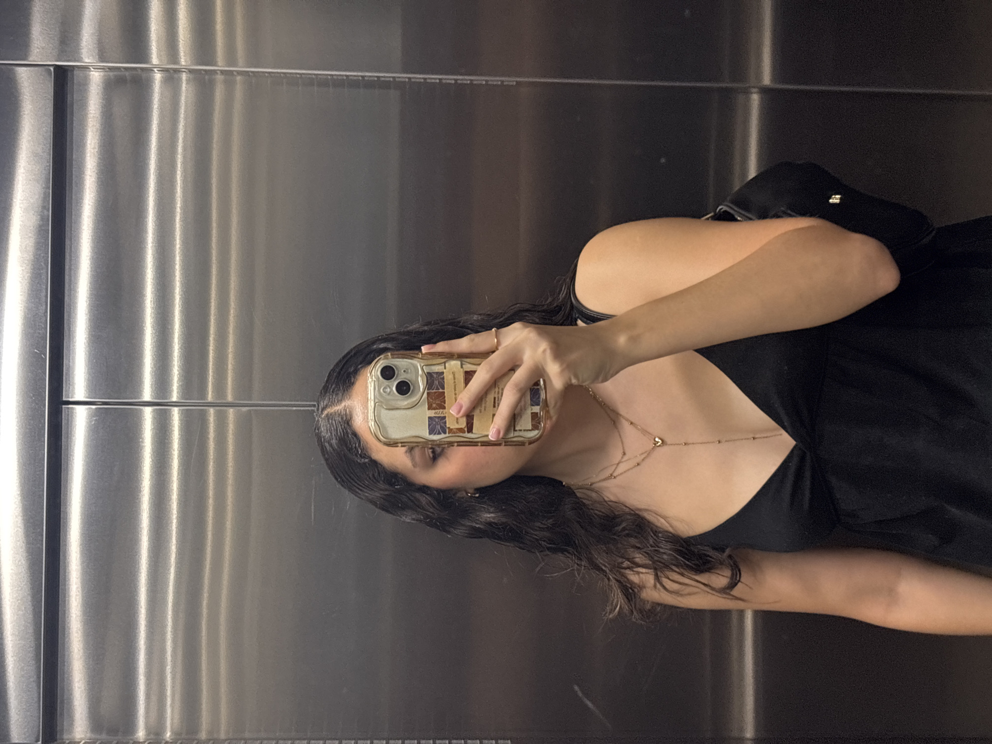 In an elevator in the city of San Andrés Cholula, Mexico, a young adult female with long, dark, wavy hair is captured in a mirror selfie. Her face is partially obscured by a smartphone held in her right hand. She wears a black, V-neck or camisole-style top. Layered gold-toned necklaces adorn her neck, one featuring a small heart pendant and a beaded drop chain, the other a shorter, simple chain. A delicate gold-toned ring is visible on her right ring finger. The smartphone, likely an iPhone, is encased in a clear cover featuring gold-toned borders and rectangular inserts with muted, patterned designs. A black shoulder bag or fanny pack with a metallic clasp detail is slung over her left side, partially visible behind her arm. The elevator interior features brushed metallic silver walls with horizontal striations and a vertical seam, reflecting ambient light.