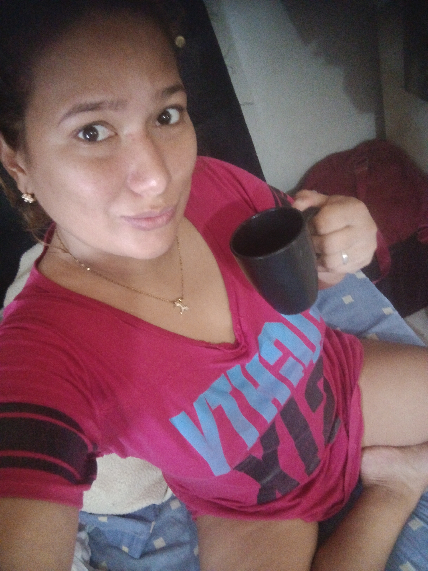 A medium-shot, selfie of a tan skinned adult woman wearing a pink v-neck short sleeved shirt with blue and dark gray text, and a gold necklace and earrings. She is holding a black mug. She appears to be in bed, with a blue patterned sheet. She has dark hair and is looking at the camera.
I lack the information to confirm the location in Caracas, Venezuela.