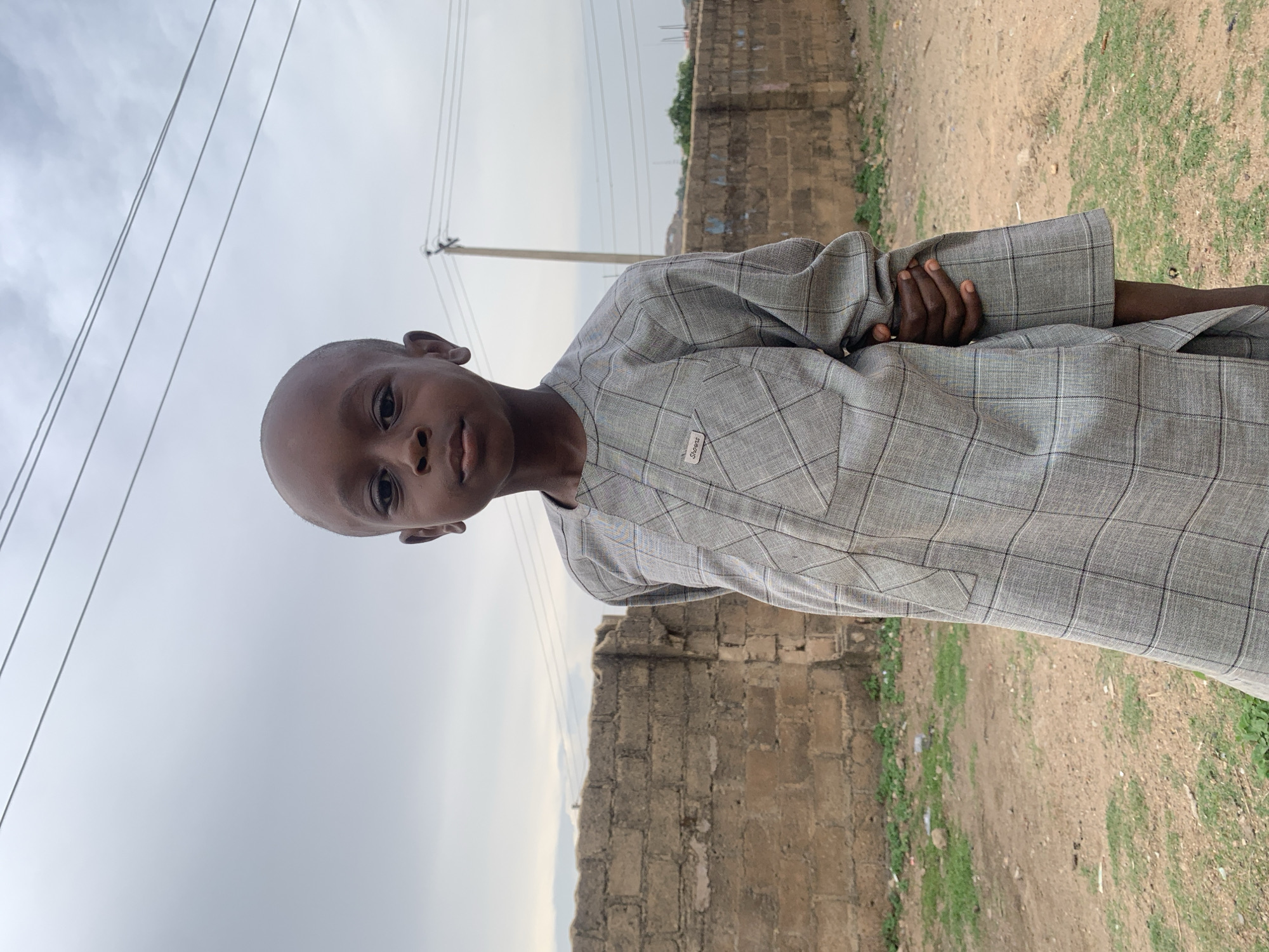 A young boy is standing outdoors. He is wearing a gray, plaid, long-sleeved shirt, and has a shaved head. He has his arms folded. Behind him is a dirt path or road, a low brick or stone wall, and some sparse green vegetation. Overhead, there are power lines against a cloudy sky. The location is Jos, Nigeria.