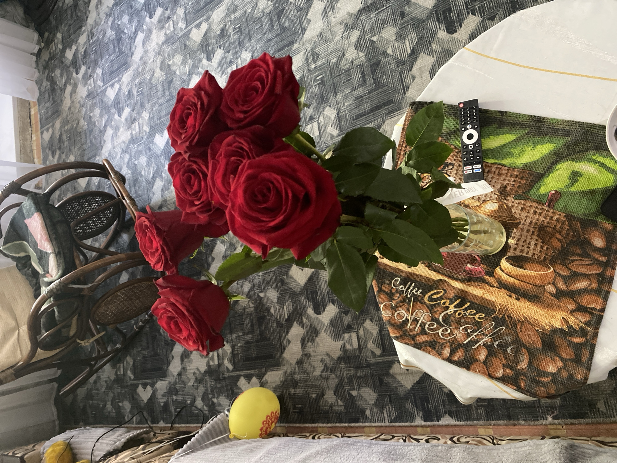 The image depicts an indoor scene, likely a living room, with a focus on a bouquet of red roses in a glass vase. The roses are arranged on a round table covered with a tablecloth featuring a coffee bean and wheat design. A black remote control rests on the tablecloth near the vase. In the background, a wicker rocking chair is visible, and a patterned carpet covers the floor. To the left, a yellow object, possibly a decorative item or a toy, hangs from a white textured surface. The lighting suggests an indoor setting with natural light possibly coming from a window, indicated by the white sheer curtains in the upper right corner. There are no people visible in the image, and no specific details about the location in Chernogorsk, Russia, can be determined from the visual information provided.