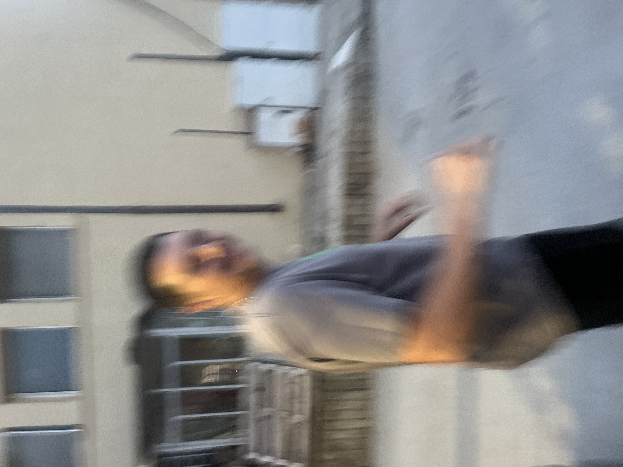 This blurry outdoor photograph, taken in Basarbovo, Bulgaria, captures a light-skinned adult male in motion, positioned right of the frame's vertical center. He is wearing a grey short-sleeved t-shirt and dark trousers/shorts, with his right arm extended forward. His facial features are heavily blurred but suggest a slight smile or expression of amusement. In the background on the left, a light-colored building with multiple rectangular windows on an upper level is visible, with a lower section featuring vertical bars or a grated structure. To the right of the subject, a plain, light-colored wall stands behind several rectangular objects resembling utility boxes or outdoor cabinets, one appearing brownish-orange and two lighter in color. The ground, likely a paved surface, exhibits strong motion blur, indicating the subject's movement within an urban or semi-urban environment.