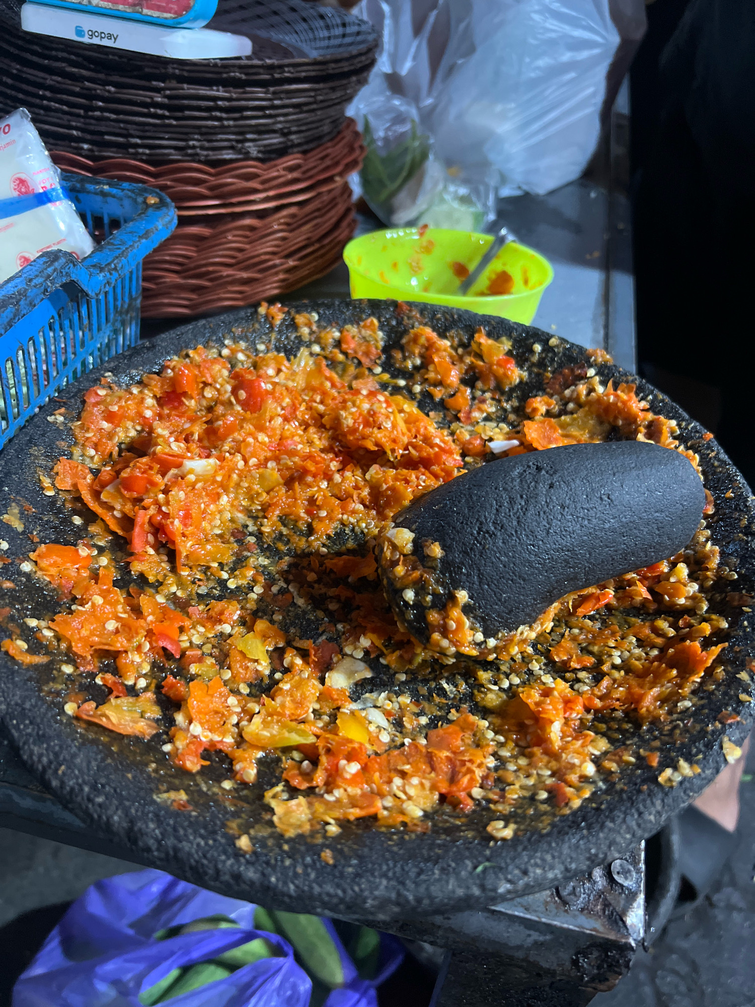 This image captures a close-up view of a traditional mortar and pestle, filled with finely chopped red chilies and other ingredients, likely being prepared as a sambal or chili paste. The mortar is made of dark, rough stone, with the pestle resting inside. The surrounding environment suggests a food stall or kitchen setting in Tlogoadi, Indonesia. In the background, stacks of woven brown baskets are visible, along with a blue plastic basket holding some packaged items. A bright green plastic bowl is also present. To the lower left, a purple plastic bag contains what appear to be cucumbers. The lighting suggests it might be nighttime or an indoor setting.