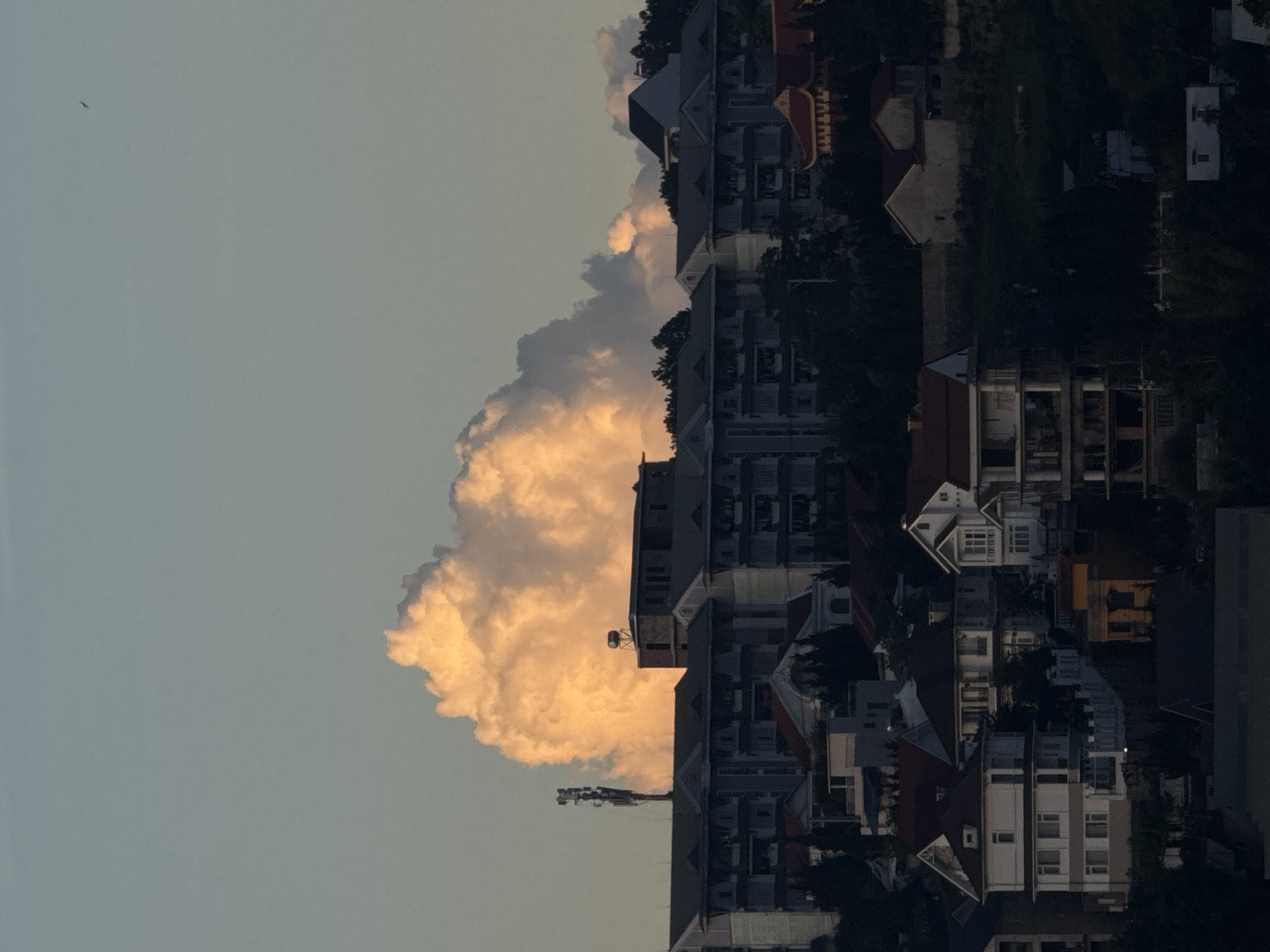 The image shows a residential area in Lang Biang - Da Lat, Vietnam under a cloudy sky. Several multi-story buildings are visible, along with houses and vegetation. The lighting suggests it might be either early morning or late afternoon. There is a large, golden-tinged cloud partially obscuring some of the buildings. An aerial is seen near the foreground. The sky is a pale blue-grey.