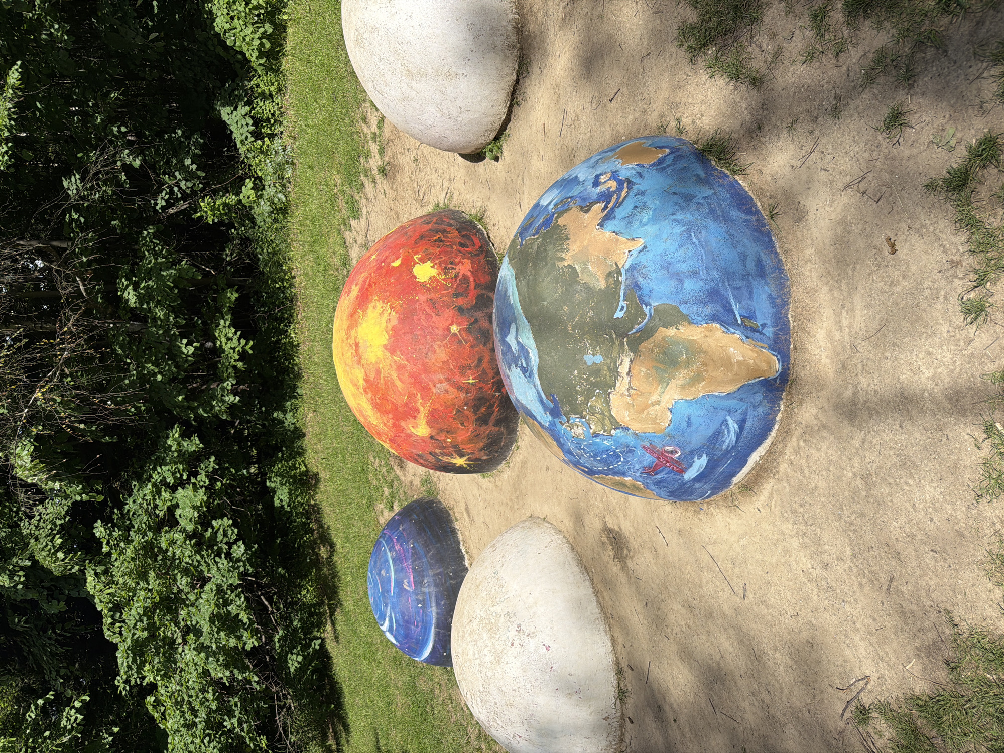 The image shows a display of painted planet models outdoors. There are at least four spherical or egg-shaped models, painted to represent different celestial bodies. One is painted to resemble Earth, with blue oceans and brown continents; a small red biplane is painted near the coastline. Another is colored red and orange, possibly representing Mars or another fiery planet. A third is blue with streaks of purple, like a gas giant, and the last one is white. The models are placed on a dirt or sand-covered ground with grass on either side. To the left of the models is a dense green bush. The setting is likely a park or outdoor educational area. The image was taken at an angle and turned; the actual location of the models is unknown. Given the model of Earth featuring a logo, it is likely located at KidZania Sofia.