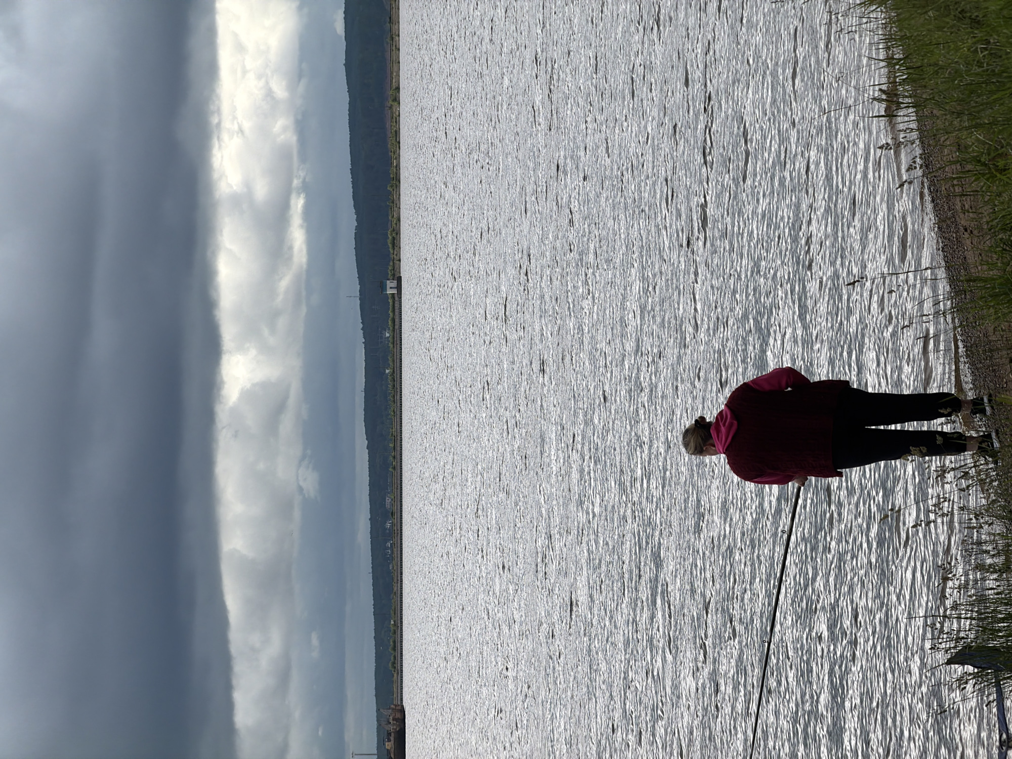A wide-angle landscape photograph captured from a slightly elevated perspective depicts an individual fishing on the bank of a large body of water under a heavily overcast sky. The location is the city of Chaikovsky, Russia.
In the foreground, an adult, likely female, is observed from behind, standing on a narrow strip of pebbled and sandy shoreline bordered by green and dried grasses. She is wearing a dark red or maroon sleeveless vest over a pink hooded sweatshirt, dark pants with a faint pattern on the lower legs, and dark athletic shoes. Her light-colored hair is tied back in a bun or ponytail. She holds a dark fishing rod, extended over the water to the left of the frame.
The expansive body of water, possibly a river or reservoir, occupies the majority of the mid-ground, displaying numerous small ripples and reflections from the sky. Across the water, a distant, dark green treeline or forested hill runs horizontally, behind which faint structures and a road are discernible. To the far left, a dark, large industrial barge or structure is partially visible along the distant bank. Further to the right along the distant shoreline, a small white building with a dark roof stands on what appears to be a pier or embankment.
The sky is dominated by dense, dark grey stratocumulus clouds, with a lighter band of clouds visible below the uppermost layer, indicating a heavily overcast day with diffused, muted light. The overall scene conveys a quiet, possibly somber, outdoor activity under an impending or persistent cloudy condition.