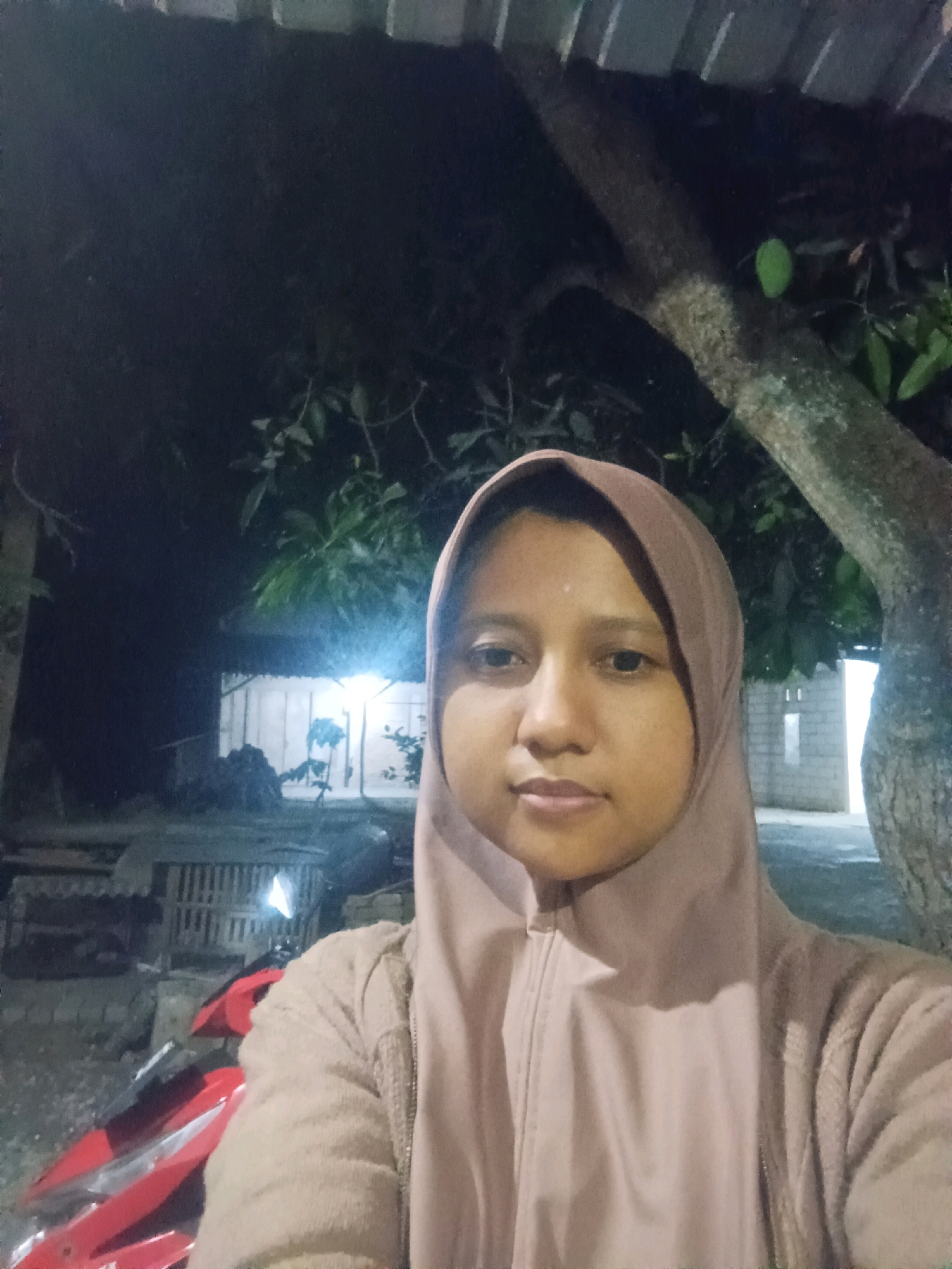 A young Indonesian woman wearing a light brown hijab and a light brown zip-up hoodie is taking a selfie outdoors at night. She is positioned in the foreground, with her face filling the majority of the frame. Behind her, a large tree trunk is visible on the right side of the image. In the background, there is a dimly lit structure with what appears to be brickwork, and a red motorcycle is partially visible on the left. The overall environment suggests a residential or rural setting in Canga'an, Indonesia, likely a backyard or a street at night.