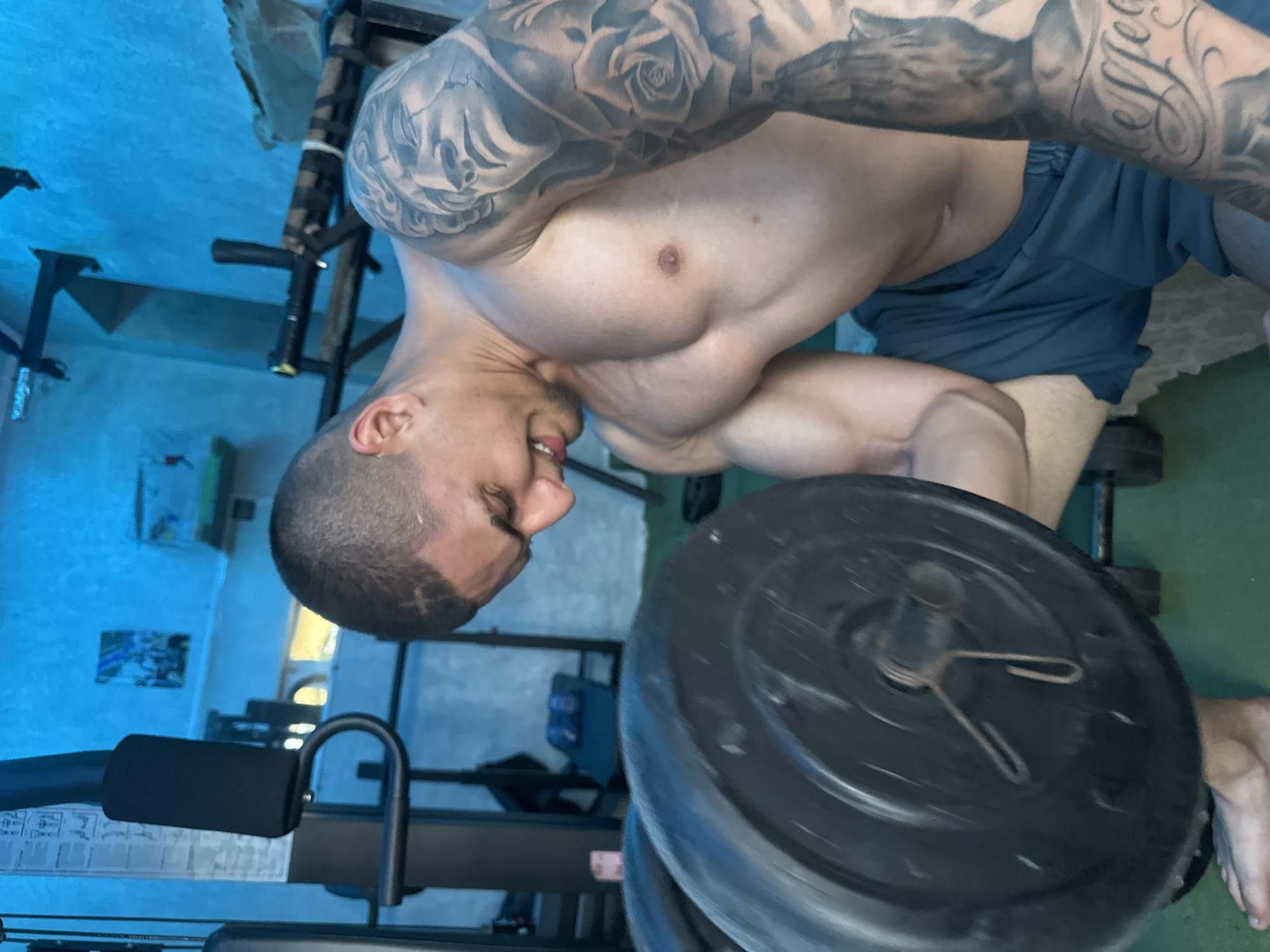 The image, oriented sideways, features a muscular man actively engaged in weightlifting. He is shirtless, showcasing a well-defined physique with prominent biceps, shoulders, and chest, and wears dark shorts. Extensive tattoos cover his upper arms and shoulders, depicting intricate roses, classical figures, and some stylized, largely illegible script. His short hair and neatly trimmed beard frame a face showing a strained expression, with his mouth slightly open, indicating exertion.
He is holding a barbell with a large, dark weight plate secured by a visible metal clip. The weight plate appears slightly blurred, possibly due to motion or shallow depth of field. The setting is an indoor space, likely a home gym, with a light-colored wall (appearing blue due to lighting) and a green floor section. In the background, various pieces of gym equipment are partially visible, including parts of a weight rack and an exercise machine with a padded handle. A black dumbbell is also seen on the floor.