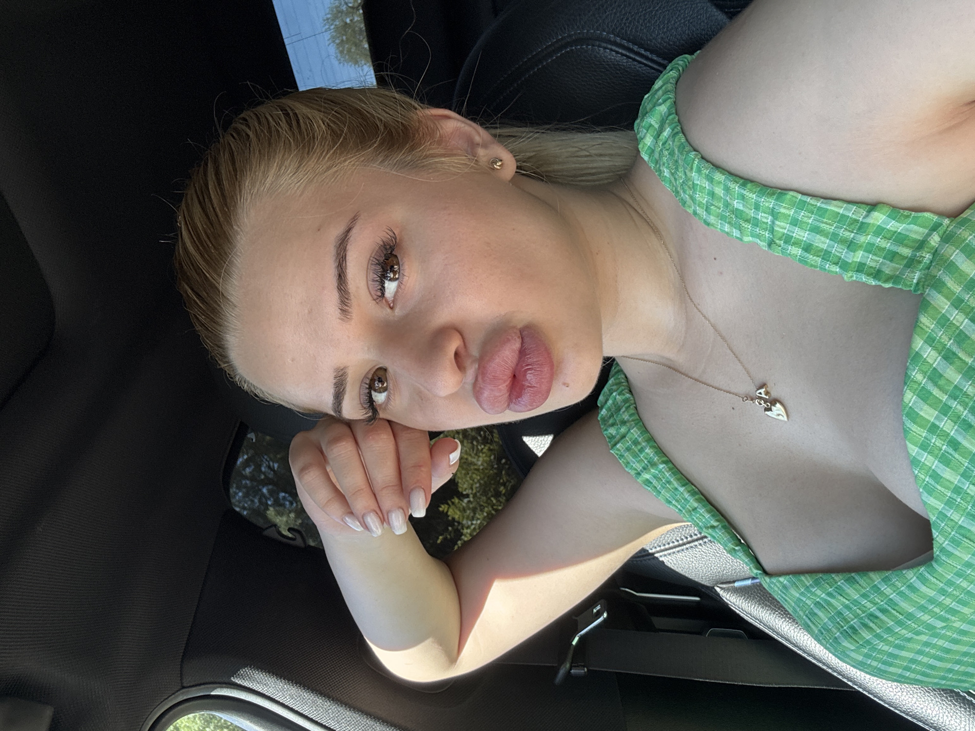 A blonde woman is in a car. She is fair-skinned and wearing a green plaid shirt and a gold necklace with a heart pendant. The interior of the car is black leather. The location appears to be Byala Slatina, Bulgaria, but this cannot be confirmed from the image alone.