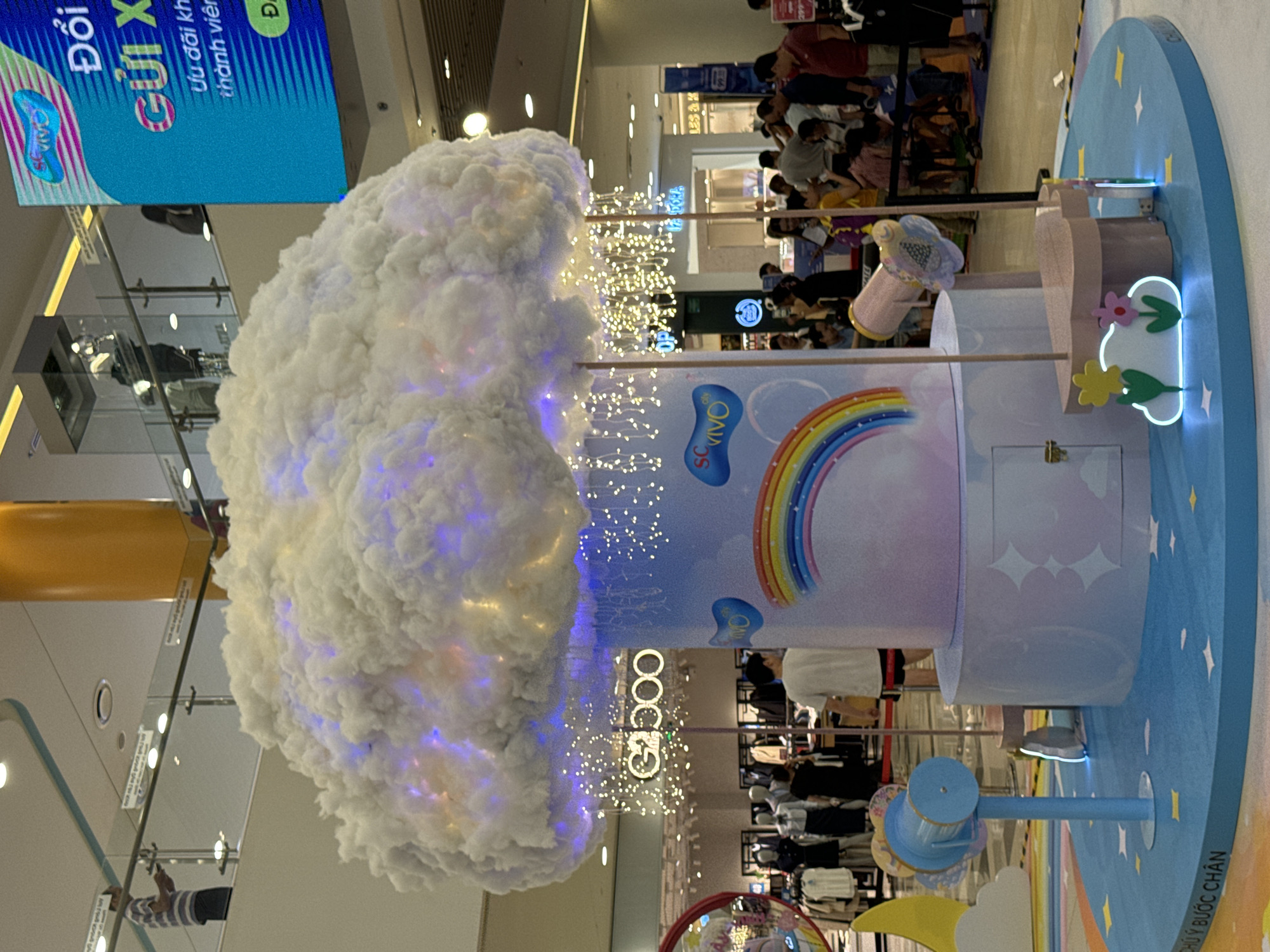 The image shows a promotional display in what appears to be a shopping mall. It's likely located in Thủ Đức, Vietnam due to the Vietnamese text in the image.
The main features include:
* A large, white cloud-like structure with blue and yellow lights.
* A cylindrical column decorated with a rainbow and the brand "SO VIVO".
* A rotating platform featuring cloud and star shapes.
* A crowd of people can be seen in the background.
* Clothing store racks are also visible in the background.
* Signage indicating promotion is available for members.
The scene depicts an indoor environment, a shopping mall during a promotional event, with people milling around.