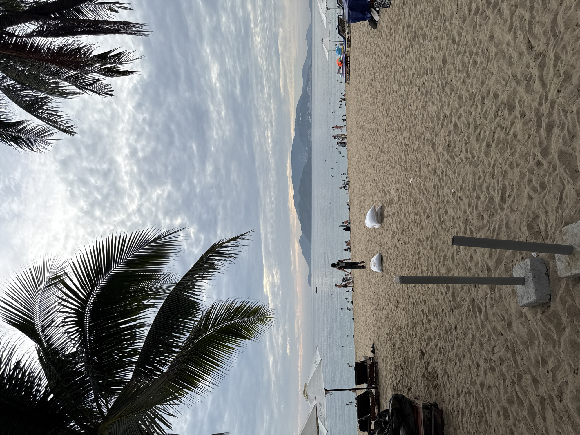 This is a medium shot of a beach in Nha Trang, Vietnam. The foreground is dominated by a sandy beach with several sandbags and two concrete posts. In the midground, people are visible walking on the beach, swimming in the ocean, and relaxing under umbrellas. The ocean stretches to the horizon, with a distant mountainous landscape under a cloudy sky. Palm trees frame the top of the image.