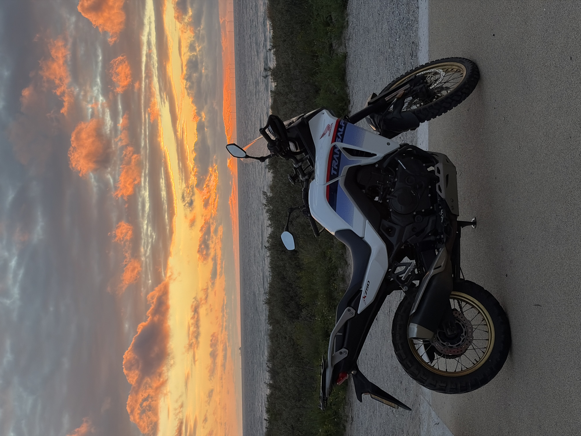 An adventure motorcycle is parked on a paved surface, with its front wheel turned slightly to the left. The motorcycle is white with blue and red decals that read "TRANSALP". The background features a body of water, likely the sea, with a low, green, grassy embankment in the foreground. Beyond the water, a row of wind turbines is visible on the horizon, suggesting a coastal location. The sky above is filled with dramatic clouds, illuminated by the warm orange and yellow hues of a sunset. The scene appears to be set at the coast near The Hague, Netherlands, given the presence of the sea and wind turbines. There are no people visible in the image. The overall impression is one of a serene, natural landscape at dusk.