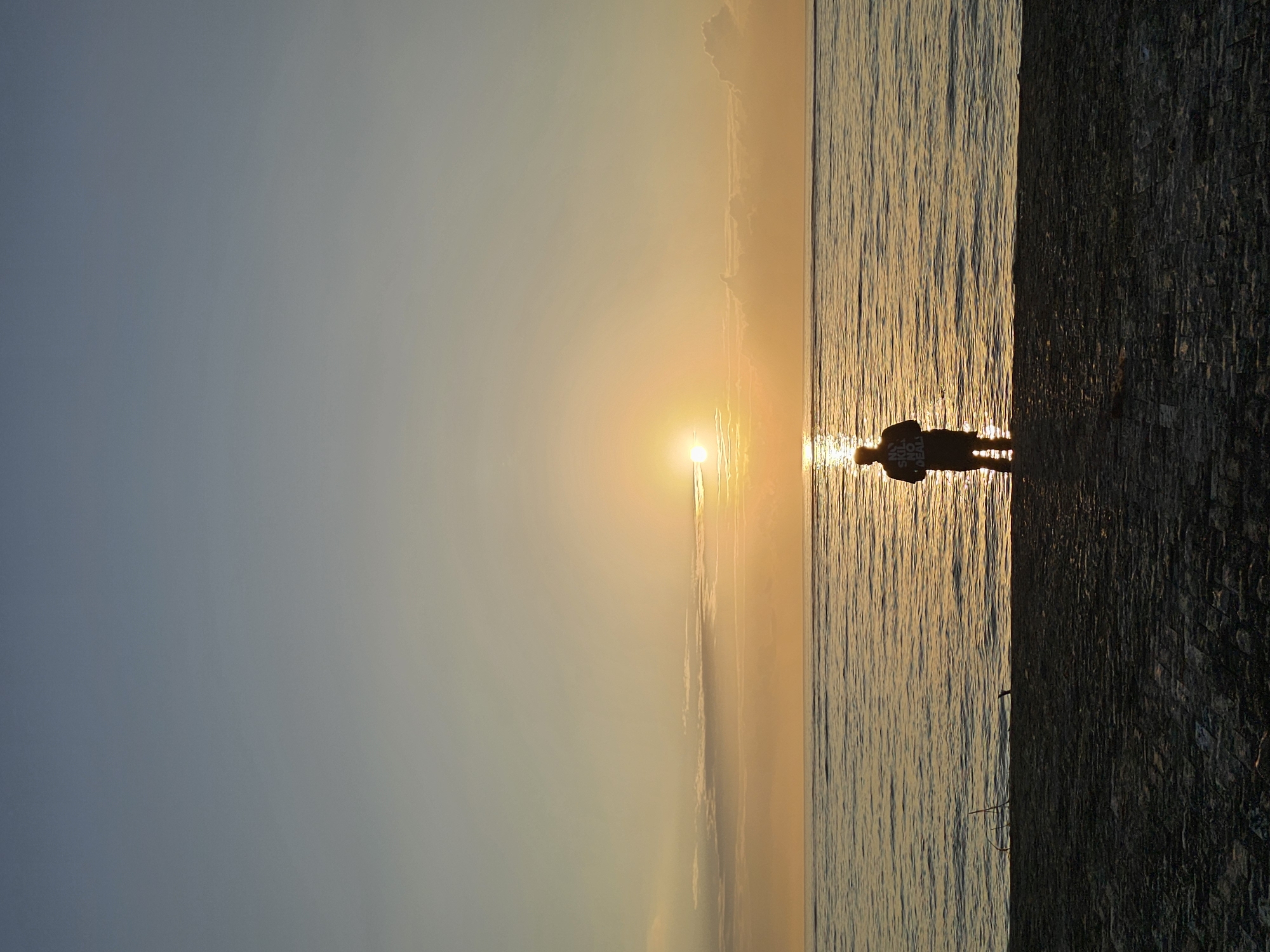 A single person stands facing a sunrise over the ocean. The person, silhouetted against the bright sun, is wearing a t-shirt and shorts and is partially submerged in the water. The sky transitions from a pale blue at the top to a warm orange and yellow near the horizon where the sun is positioned. Wispy clouds are visible on the horizon. The ocean is calm with gentle waves reflecting the sunlight. The foreground shows a dark, textured surface, possibly a beach or walkway, with some debris. The image appears to be taken from a low angle. The location is unspecified, but the overall scene suggests a coastal setting at sunrise.