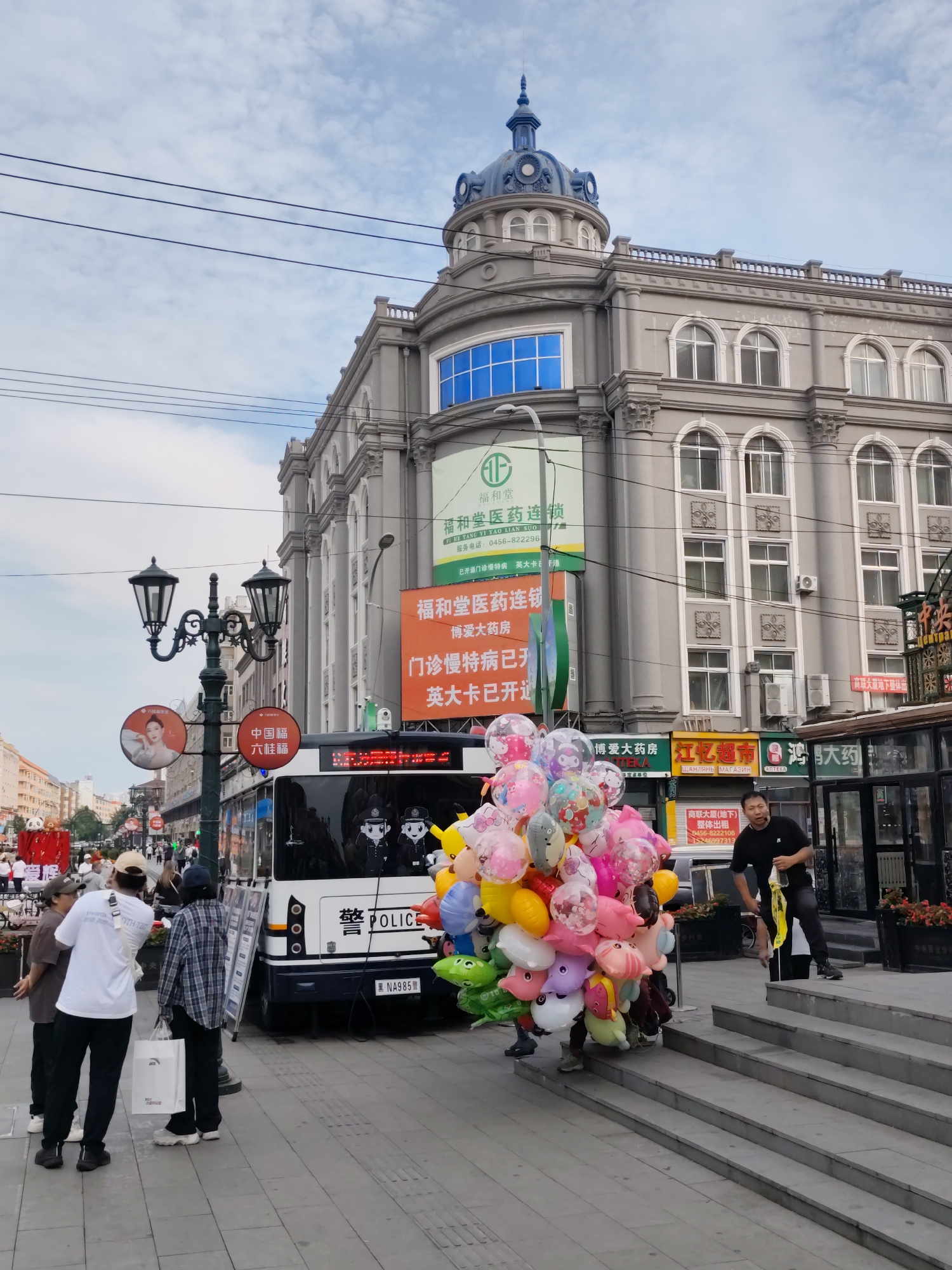 In the city of Xingan Subdistrict, China, a street scene unfolds with a large, ornate building dominating the background. The building, with a distinctive blue dome and classical architectural elements, houses various businesses, including pharmacies. In the foreground, a street vendor is seen selling a large cluster of colorful balloons. He is dressed in a black t-shirt and dark pants, and is holding a yellow string. Several people are gathered on the sidewalk, including a group of three individuals wearing casual clothing and a person in a plaid shirt. A white bus, marked with "POLICE" and Chinese characters, is parked nearby. The street is paved with gray tiles, and a lamppost with multiple lights stands prominently. Overhead, electrical wires crisscross the sky, which is partly cloudy. The overall atmosphere suggests a bustling urban environment.