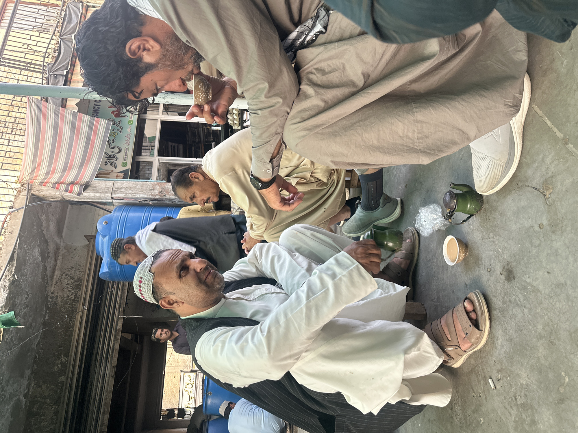 In Quetta City Tehsil, Pakistan, a group of men is gathered in an urban setting. One man, in a light-colored outfit, sits while another man in a gray outfit appears to be examining or smelling something held close to his face. A smaller, light-colored tea cup rests on the concrete floor near an old green teapot. The scene unfolds in an outdoor area, where blue water storage tanks and various other objects create a sense of being in a public space. The building façade is visible in the background, complete with signage and awnings.