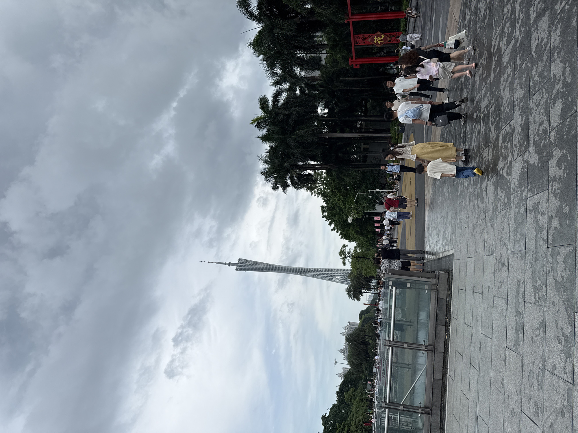 The image depicts a view of the Canton Tower, a prominent landmark in Guangzhou, China, located in Tianhe District. The tower, a slender, twisting structure, dominates the skyline under a cloudy sky. In the foreground, a paved walkway is populated by a diverse group of people. Some are walking, while others appear to be standing and observing. Several individuals are dressed in casual attire, including t-shirts, shorts, and skirts. Among the crowd, there is a young boy walking ahead of an adult woman, and a couple walking together. Palm trees and lush green foliage frame the scene, with some buildings visible in the distance. A red archway with Chinese characters is also present in the background. The ground surface is wet, suggesting recent rain.
