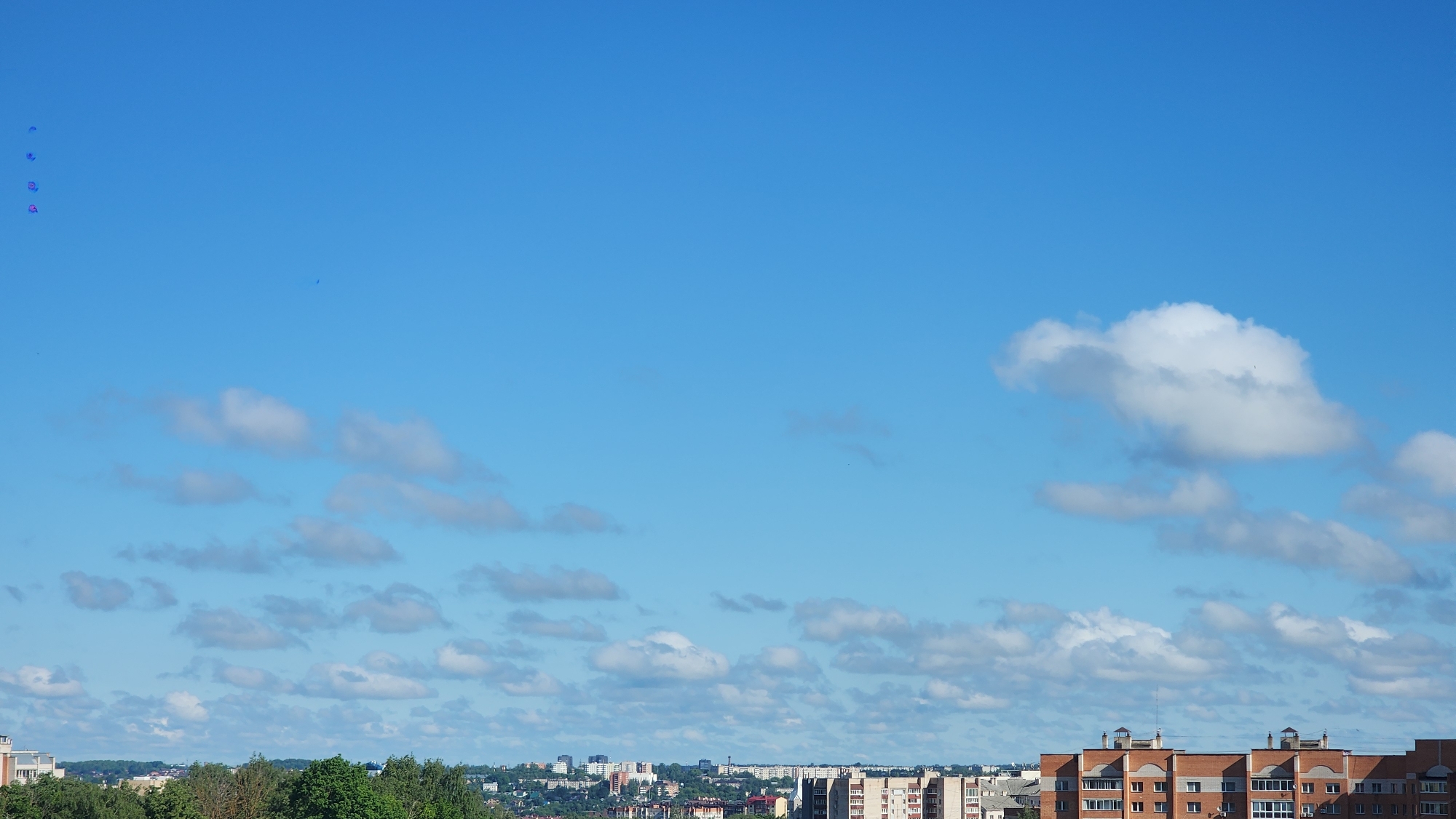 The image captures a daytime view of the city of Smolensk, Russia, under a blue sky with scattered white clouds. The foreground consists of buildings and green treetops. A mix of residential and possibly commercial buildings are visible, varying in height and architectural style. A cluster of trees separates these buildings. The middle ground includes city infrastructure stretching to the horizon. The sky dominates a large portion of the frame, showcasing a blue gradient and varying cloud formations.
