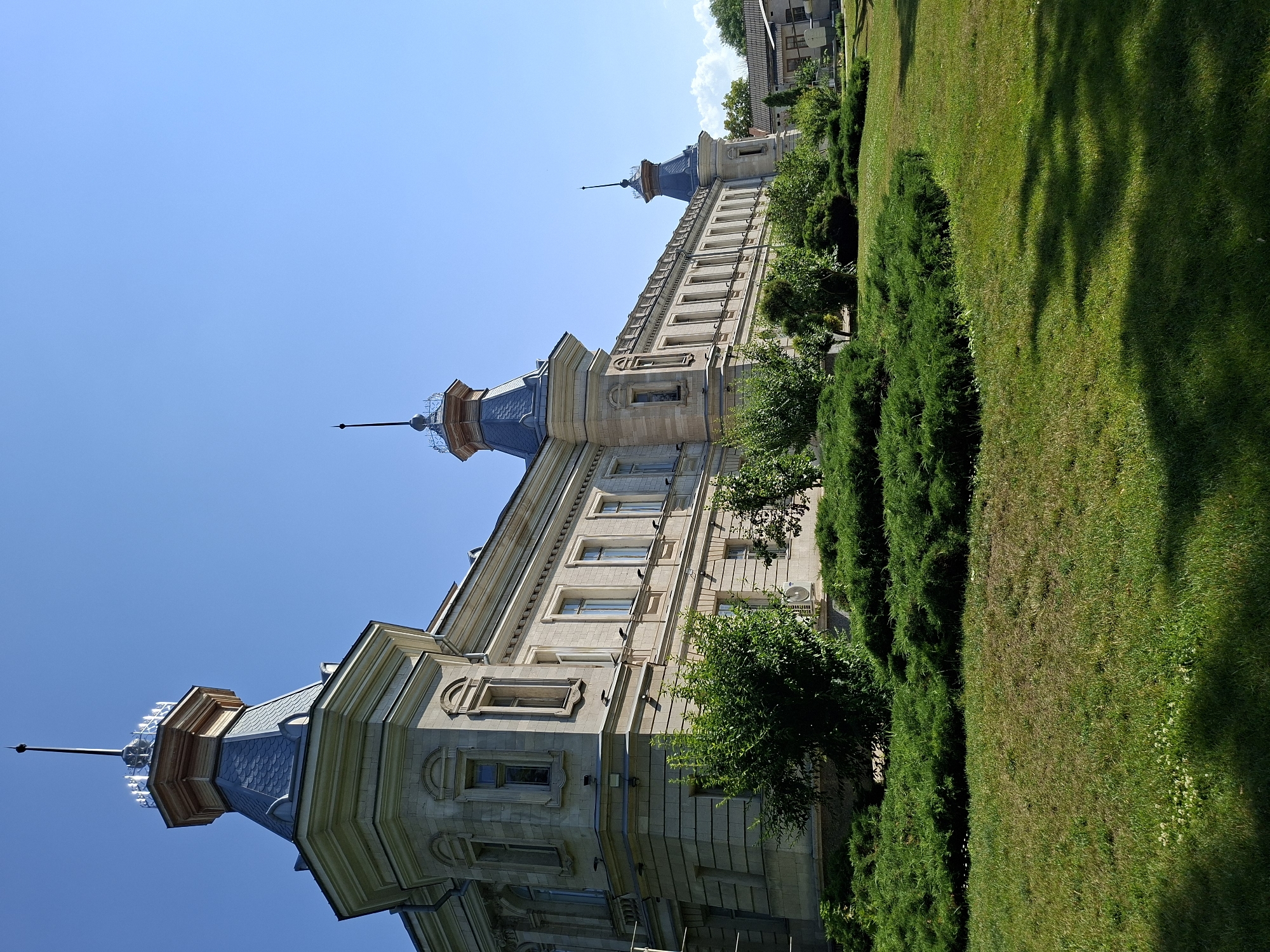 The image shows a large, ornate, light-colored stone building with multiple towers topped with spires, set against a clear blue sky. The building features classical architectural elements, including arched windows and decorative moldings. In front of the building, there is a well-maintained lawn with lush green bushes and trees. The overall scene suggests a sunny day in a park-like setting. The building is the former Archbishop's Residence in Chișinău, Moldova, now housing the Museum of the City of Chișinău.