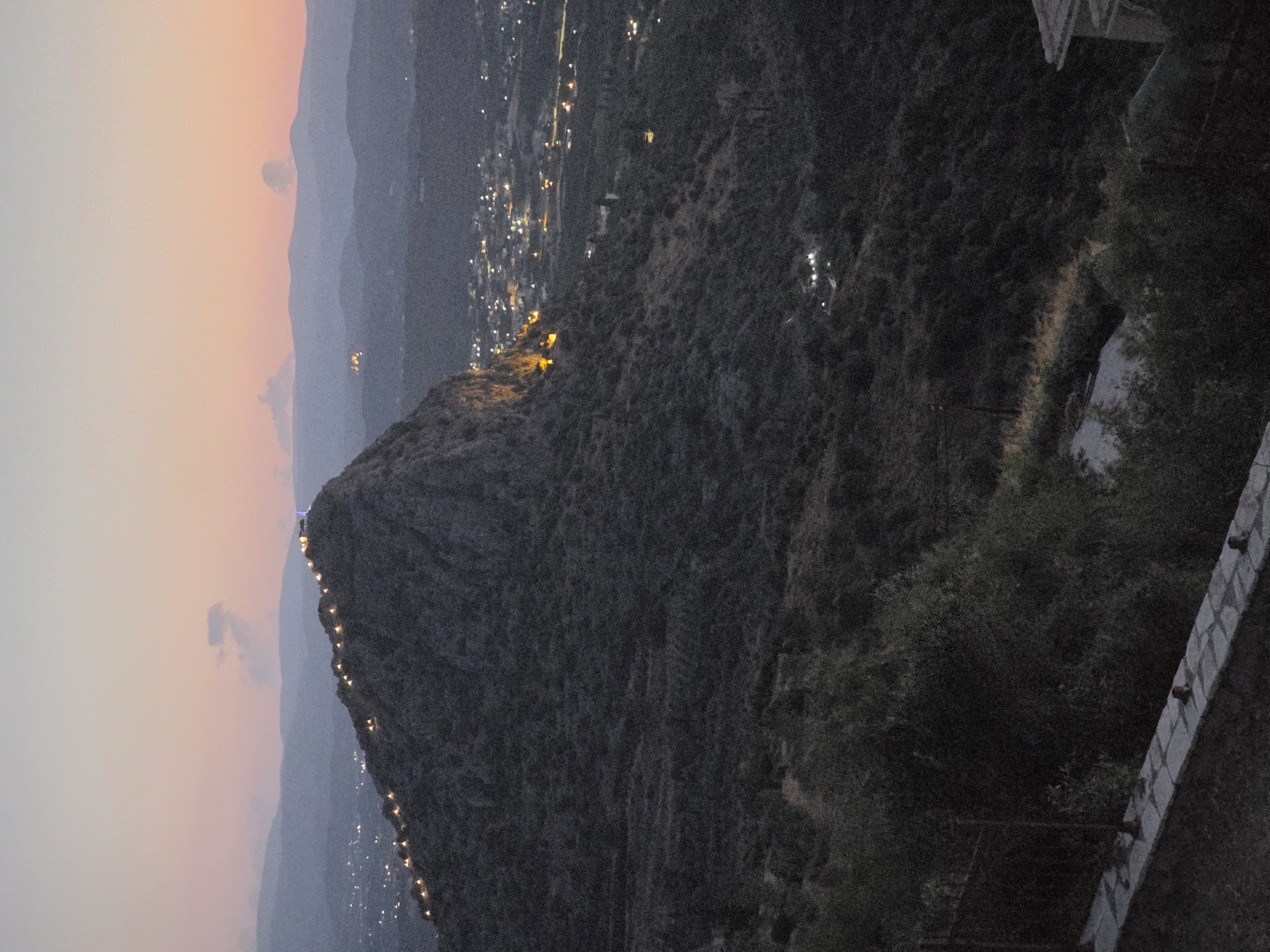 The image shows a landscape scene at dusk in the Municipal Unit of Asini, Greece. A steep, rocky mountain dominates the foreground, its peak illuminated by a string of lights. The lights appear to be decorative and follow the contour of the summit. Below the mountain, a town or village is visible, with numerous lights twinkling in the distance, indicating a settlement. The sky above is a soft gradient of pink and orange, suggesting the last moments of daylight. The surrounding terrain is rugged and covered in vegetation, with darker, dense foliage in the lower parts of the image. A low stone wall with a fence is present in the bottom left corner. The overall impression is one of a serene evening view of a town nestled near a mountainous area.