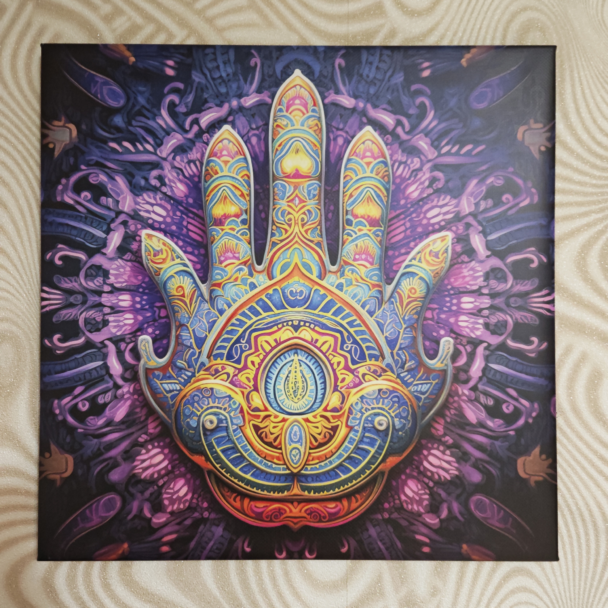 A square artwork featuring a large, stylized hand in the center dominates the scene. The hand, likely a Hamsa, is intricately decorated with patterns and an eye-like symbol. Colors include shades of blue, yellow, red, and orange. The background is a swirling, psychedelic mix of purple and black. The artwork rests against a wall with a subtle, swirling pattern. There is no evidence to suggest this image is located in the city of Rostov, Russia.