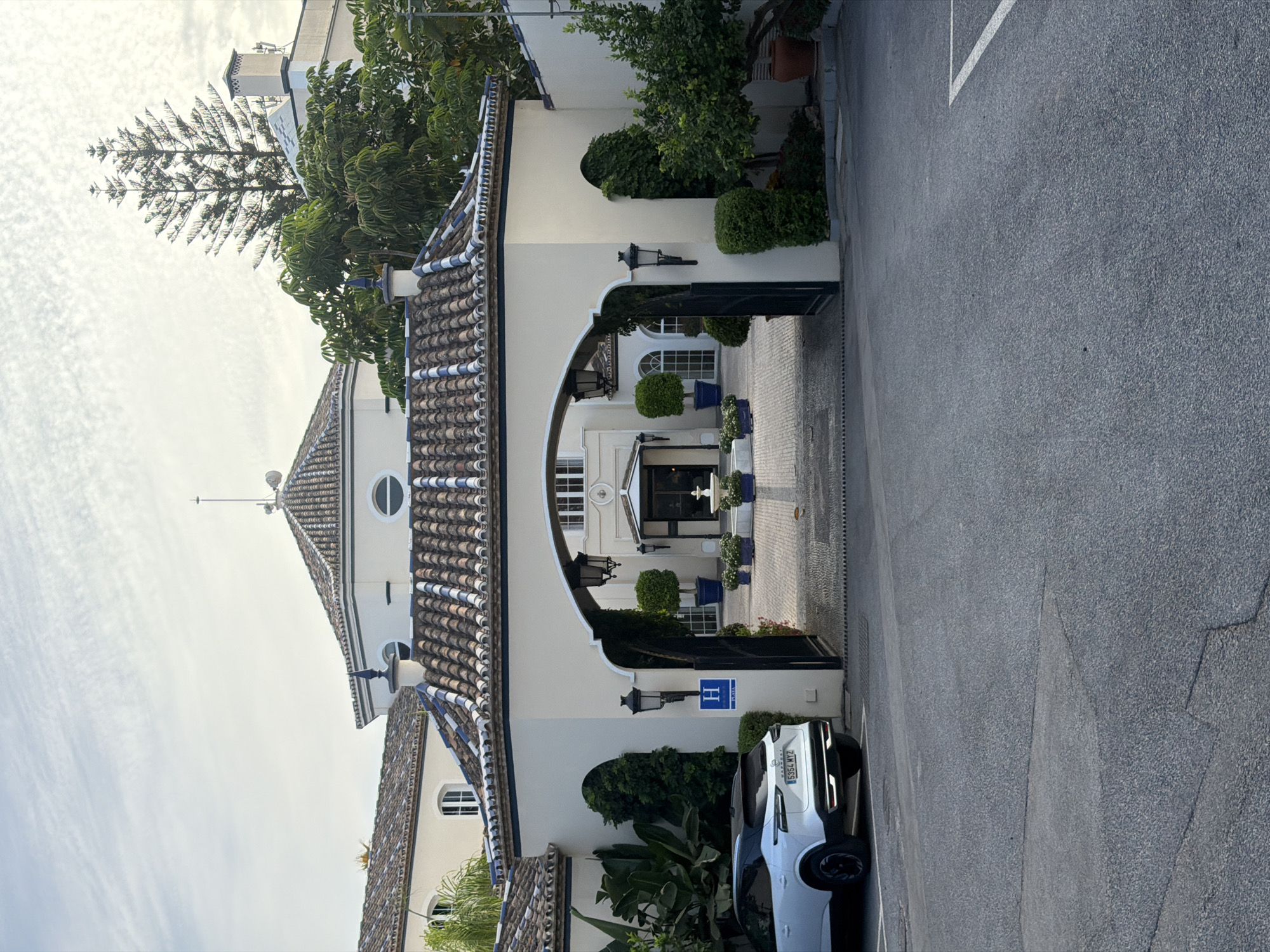 A white SUV is parked in a lot in front of an ornate entrance to a hotel. The hotel features a light-colored facade, a tiled roof with blue accents, and an arched entryway. A blue sign with a white "H" is visible on the wall to the left of the entrance. The entrance leads to a courtyard with a fountain and potted plants. Trees and lush greenery surround the building. The sky is overcast with wispy clouds.