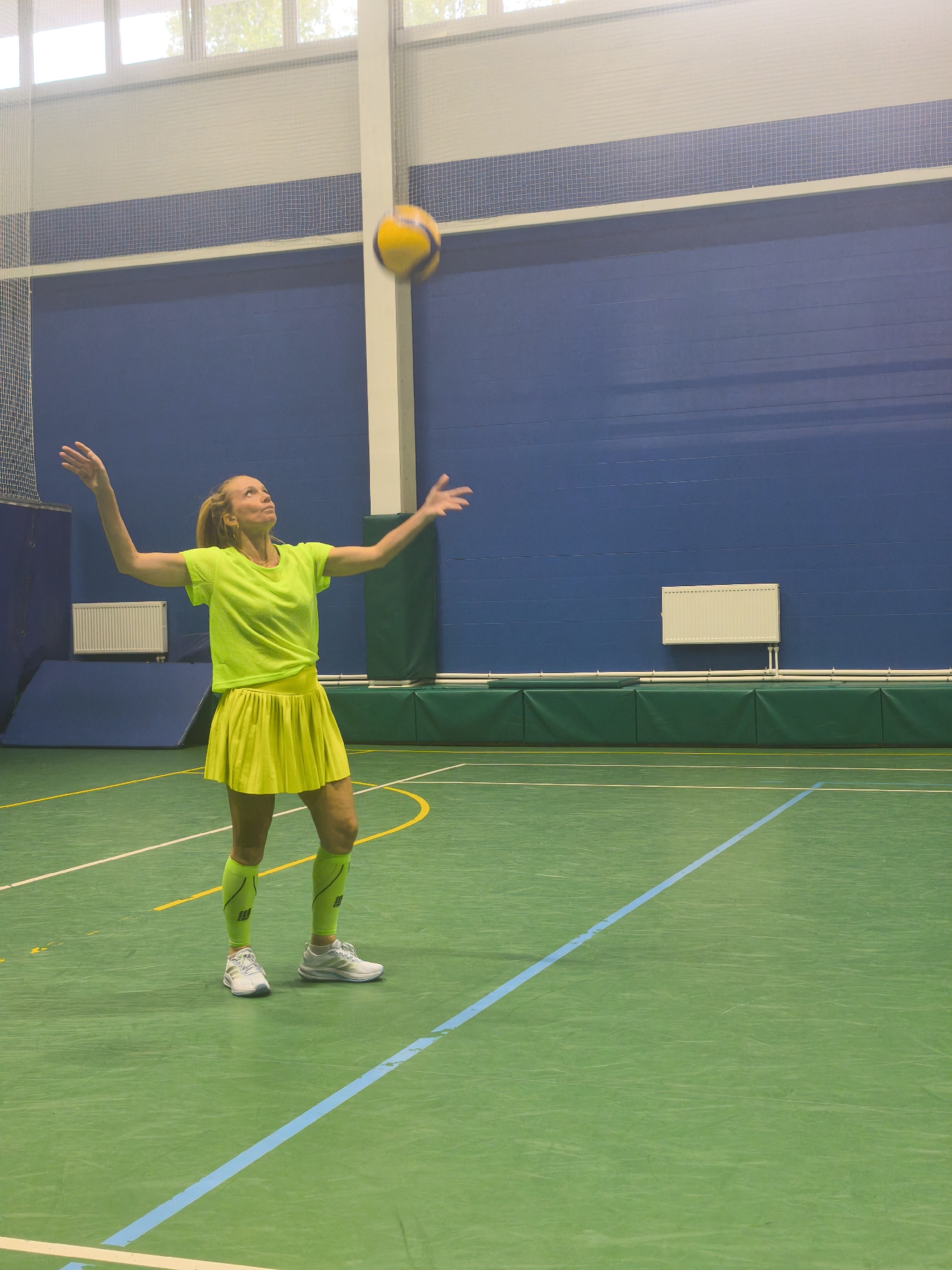 A woman in a neon yellow tennis outfit is standing on a green indoor court with blue lines, in an athletic hall. She has her arms raised and is looking up at a yellow volleyball in the air. Behind her is a blue wall with a green bottom border, and above that, a blue-tiled section and white ceiling with windows. The location of the scene is likely an indoor sports facility; however, there is no indication to conclude this image was taken in Saint Petersburg, Russia.