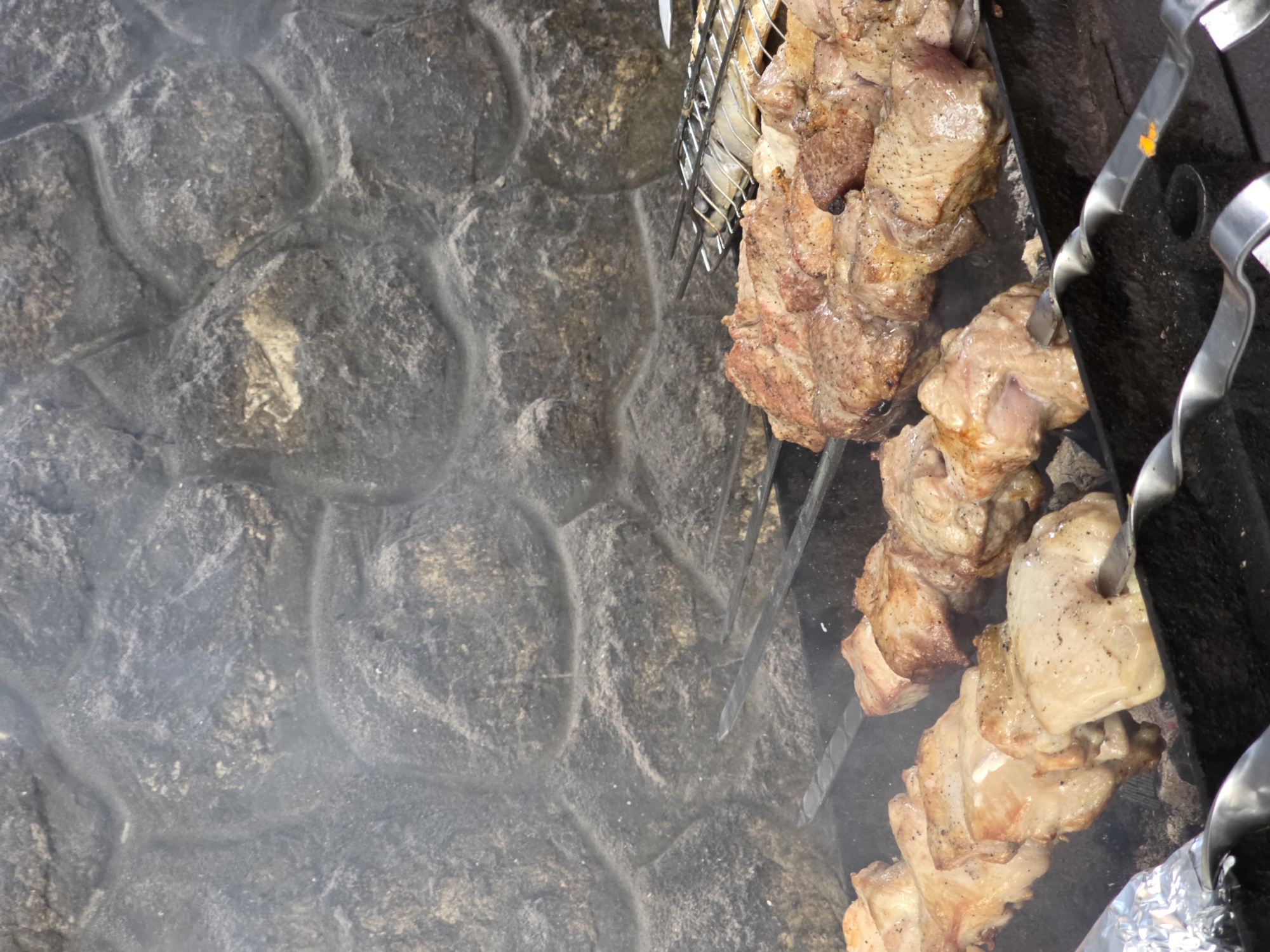 In Pidhorodne, Ukraine, an outdoor barbecue is underway, with several skewers of meat cooking over hot coals. The meat, appearing to be pork or chicken, is arranged in chunks on long metal skewers. The background features a textured stone wall, likely part of an outdoor cooking area or structure. Smoke rises from the grill, indicating the cooking process is in progress. A metal grill rack is visible in the upper right portion of the image, suggesting other items might also be cooking. The overall scene depicts a common method of food preparation in an outdoor setting.