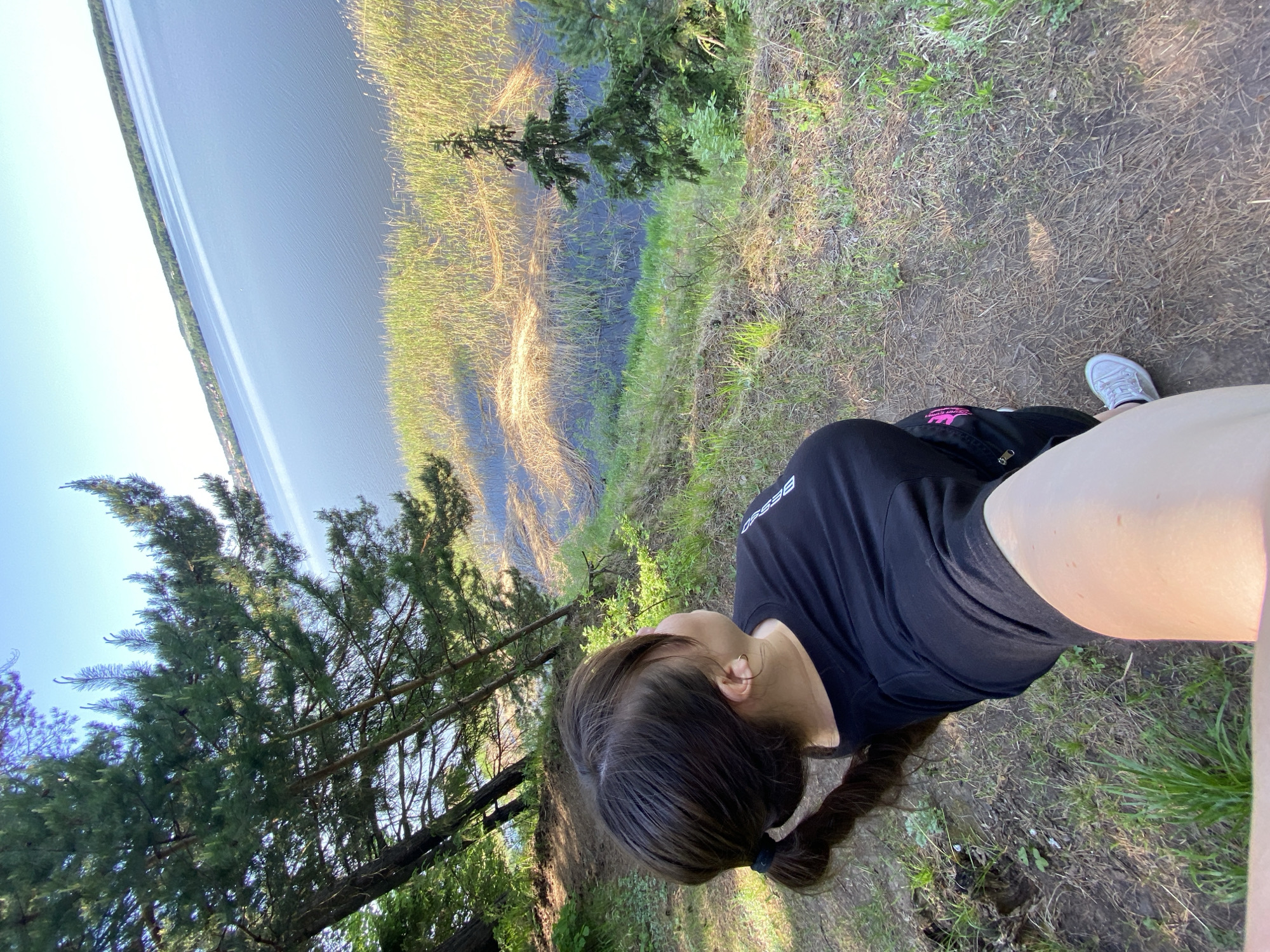 A woman stands on a grassy, slightly inclined area with trees, overlooking a large body of water. The water has a shoreline with reeds or tall grasses growing along the edge. The woman wears a black t-shirt, shorts, and light-colored sneakers. Her hair is dark and pulled back into a ponytail. Based on the environment and information provided, the location is likely an elevated area or bluff overlooking a body of water in Votkinsk, Russia.