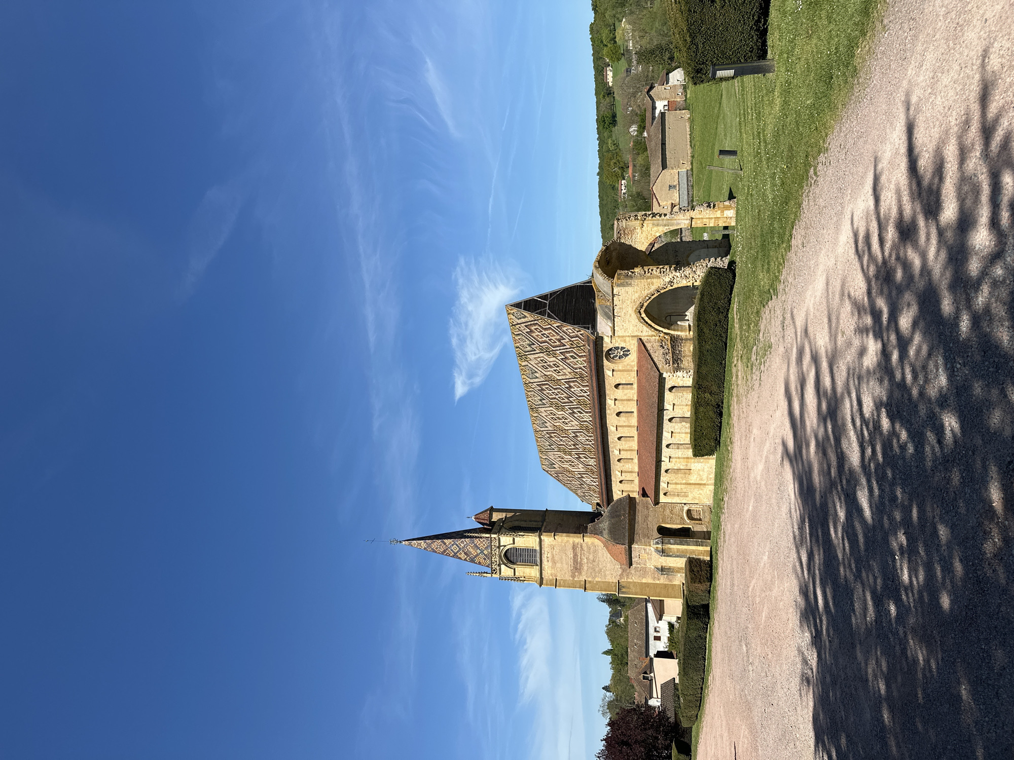 The image captures the historic Abbaye de Charlieu located in Briennon, France. The abbey is seen from a low angle, emphasizing its height against a clear blue sky with sparse cirrus clouds. The building's architecture is highlighted, featuring its iconic patterned roof tiles, a tall spire, and visible signs of age and partial ruin, particularly in the section with arched openings. A gravel path lined with manicured hedges runs alongside the abbey, marked with tree shadows. In the background, glimpses of the surrounding area of Briennon are visible, including green fields and residential buildings.