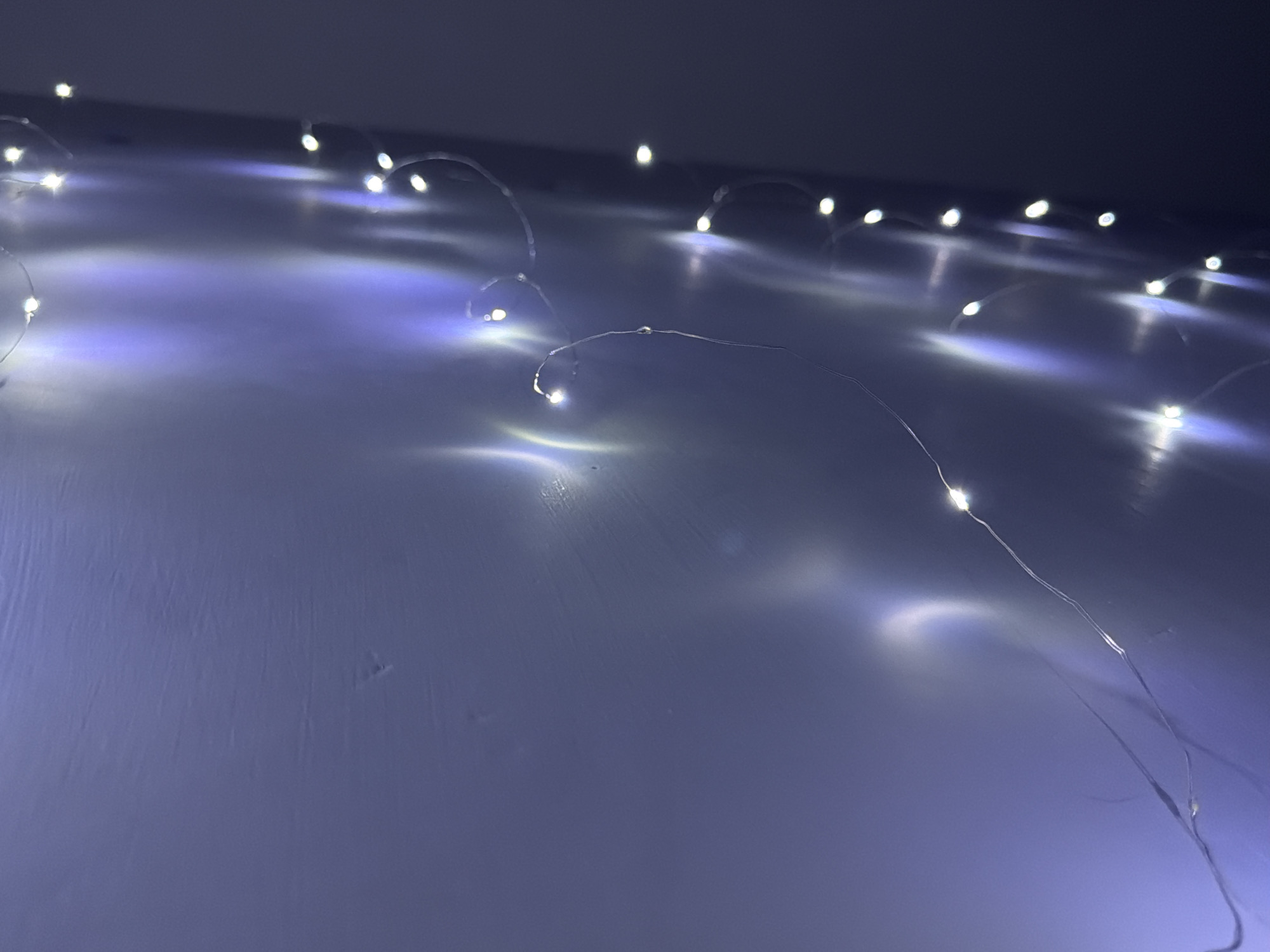 The image shows a string of fairy lights arranged on a surface. The lights emit a bright, white glow, and their reflections create a soft, blurred effect on the surface beneath. The background is dark and indistinct. The surface appears to be a solid, light-colored material, possibly a table or wall. The image was not taken in Lesnovo, Bulgaria. It is a close-up of decorative lighting.