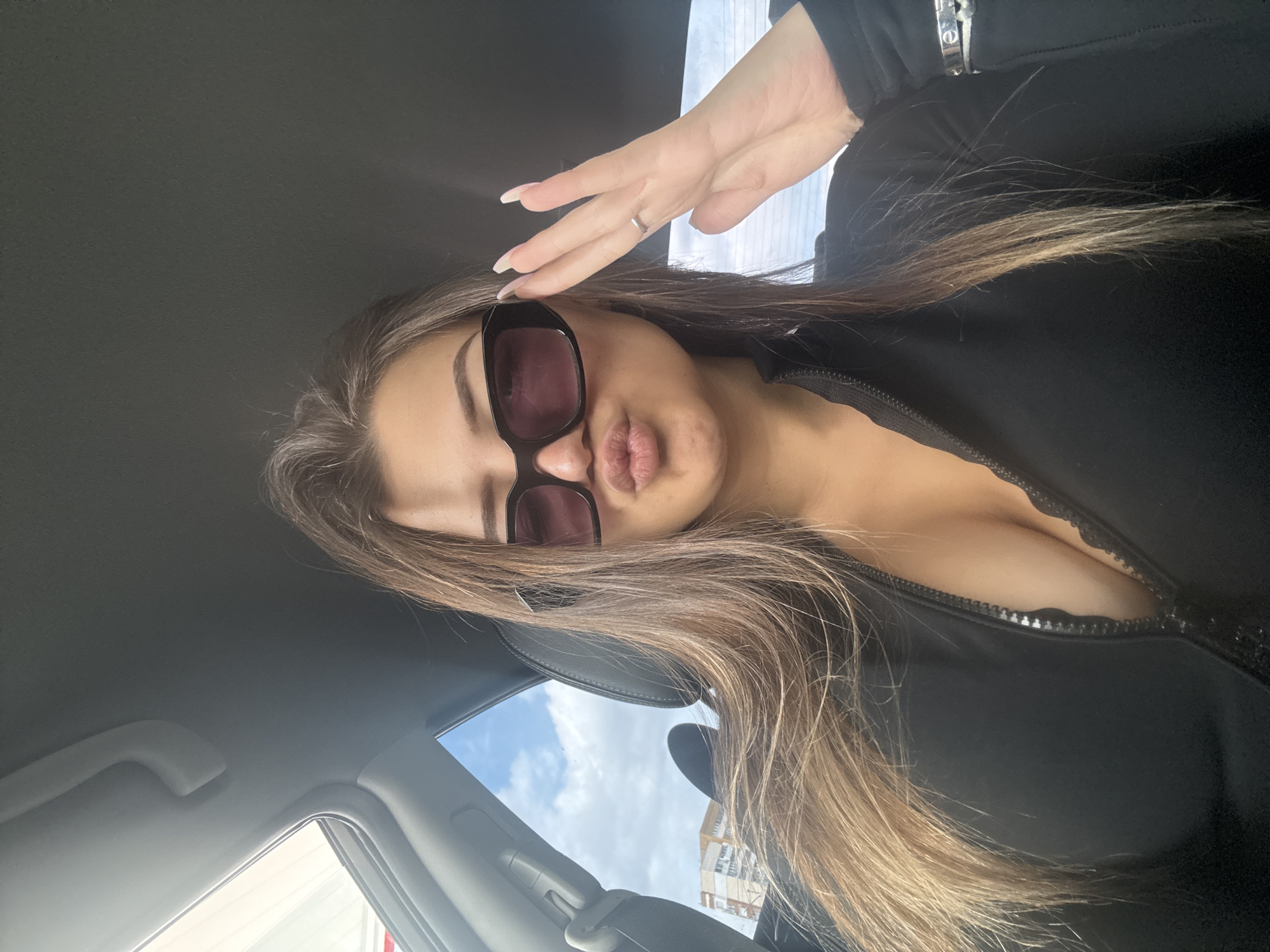A young woman with light brown hair and sunglasses is in a car. She is wearing a black zippered top. Her hand is raised near her forehead and her lips are pursed. Through the car window, there is a view of a building and sky. This image could be from Sofia, Bulgaria.