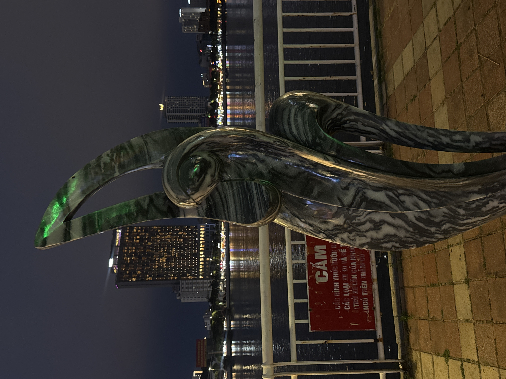 Here is a description of the image:
The image shows a marble statue of a stylized wave with a city skyline in the background, presumably in Đà Nẵng, Vietnam. The statue is predominantly gray with white veining, positioned near a metal railing. Behind the railing is a body of water reflecting the city lights. A red sign with white text, written in Vietnamese, is visible. The foreground shows a paved area with a brick-like pattern. The overall lighting suggests that the photo was taken at night.