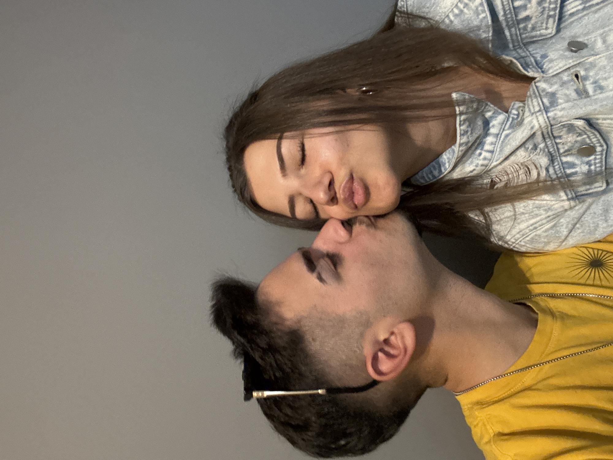 A photograph depicts a light-skinned male and female, appearing to be a couple, positioned in what seems to be a staged, intimate interaction. The woman, wearing a light blue denim jacket, is above the man, her eyes closed, lips pursed as if attempting to kiss him. The man, in a yellow T-shirt and what may be gold-rimmed glasses, has his eyes closed as well, their faces in close proximity. The background is a plain grey. No specific location details related to Sofia, Bulgaria, are apparent.