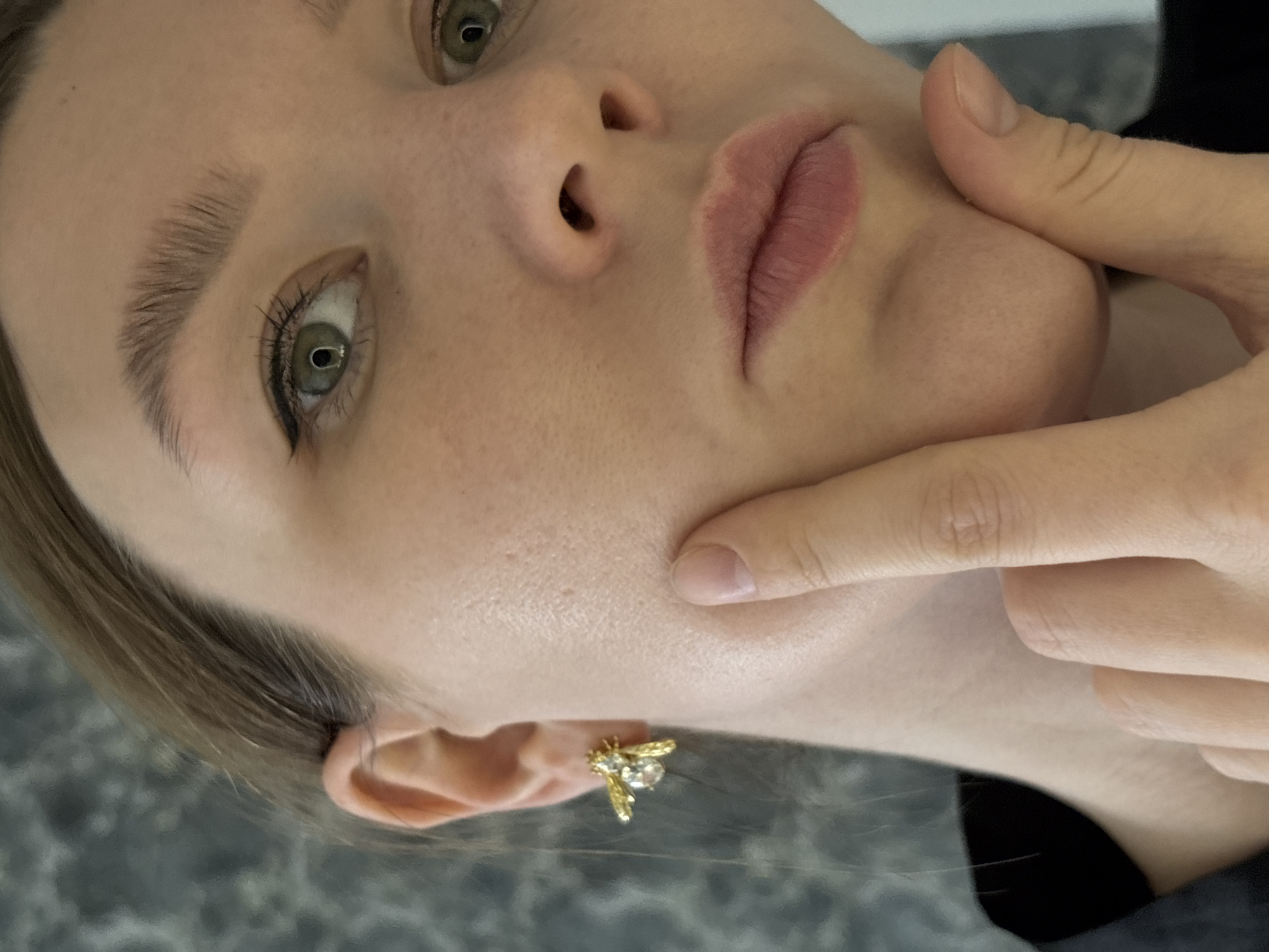 A close-up, angled image of a person's face, likely female. The person has light skin, green eyes with black eyeliner, and light brown eyebrows. One hand touches their cheek. The person wears a gold, bee-shaped earring. The background is blurry and suggests a grey surface. There is no information in the image to indicate a location in Izhevsk, Russia.