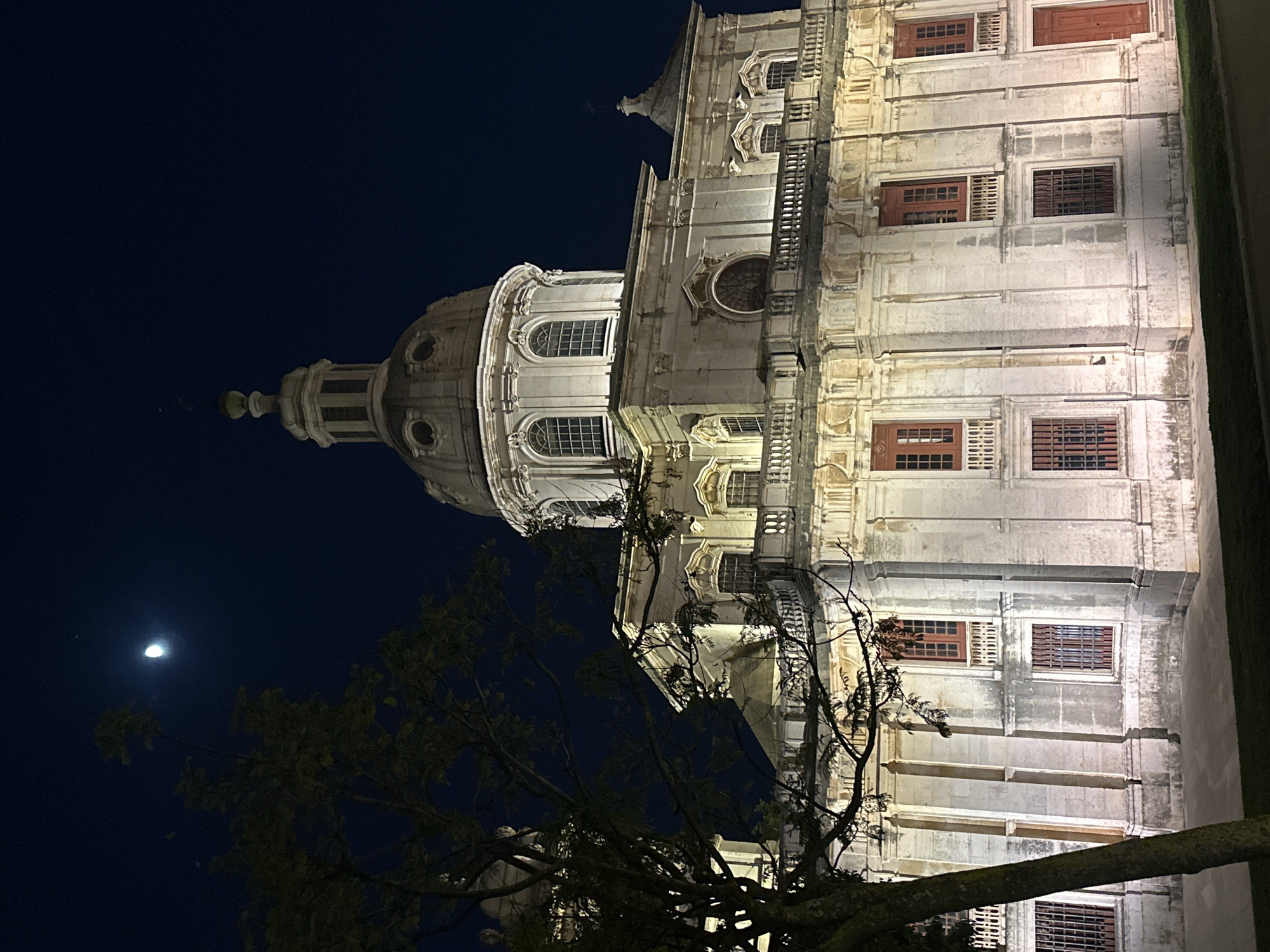 The image captures a nighttime view of the dome and facade of a grand, illuminated stone building, likely a church or cathedral, in Lisbon, Portugal. The architecture is ornate, featuring multiple windows with dark shutters, decorative moldings, and arched openings. The building's stone structure shows signs of age, with some discoloration. A portion of a tree with bare branches is in the foreground, partially obscuring the view of the building. In the deep blue night sky, a crescent moon is visible, along with a small, bright object, possibly a star or planet. The overall mood is serene and majestic, enhanced by the dramatic lighting of the architecture against the dark sky. No people or text are visible in the image.