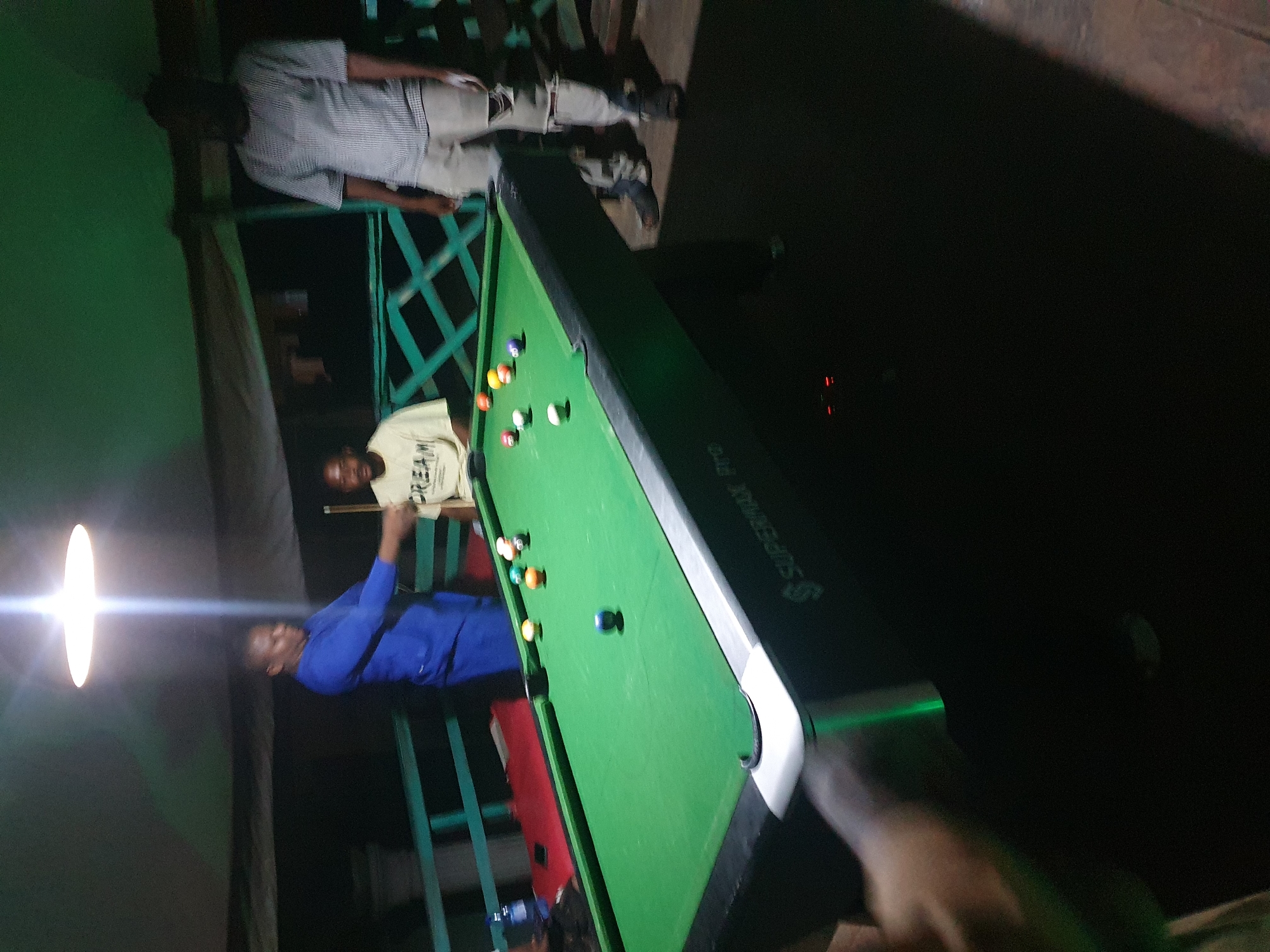 The image depicts a group of people playing pool at night, likely in Lokoja, Nigeria. Three men are visible: one in a blue shirt is actively playing, another in a light-colored t-shirt with "DREAM" printed on it is seated, and a third man in a checkered shirt and ripped jeans stands to the right, observing. A pool table with several colored balls is the central object of focus. The environment appears to be an outdoor or semi-outdoor space with a ceiling overhead and what looks like a railing in the background, suggesting a recreational area or bar. The lighting is dim, with a bright overhead light illuminating the playing area.