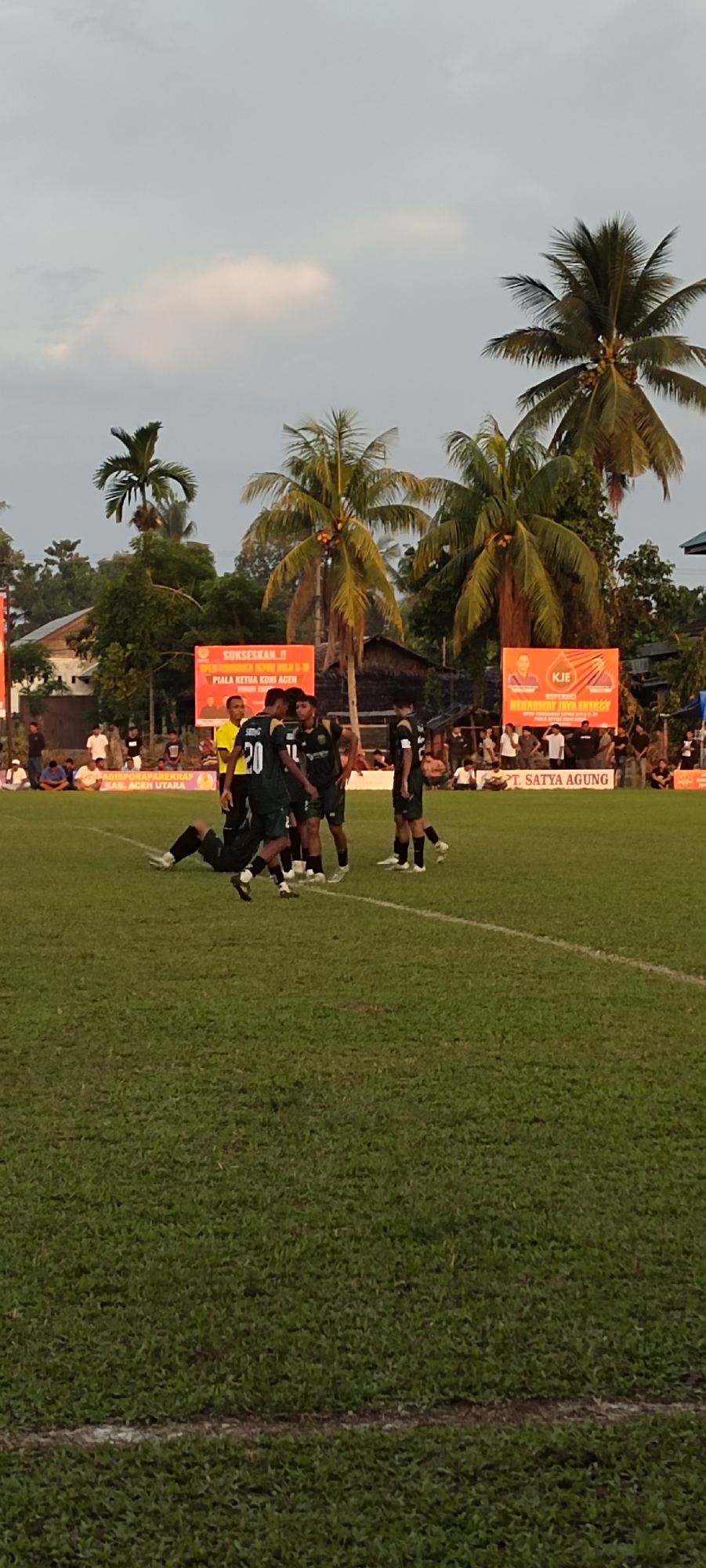 In Kilometer Delapan, Indonesia, a soccer match is underway on a grass field under a cloudy sky. The players, wearing dark green uniforms with white accents, are clustered near the center of the field, with one player appearing to be falling or lying on the ground. A referee in a yellow shirt stands nearby. In the background, palm trees and buildings line the edge of the field. Two large orange banners are prominently displayed behind the spectators. One banner reads "SUKSESKAN !! PENTINGNYA KULIAH U-18 PIALA KETUA KONI ACEH". The other banner features the logo "KJE" and text that includes "NEURUMAH JAYA ENERGY" and "PT. SATYA AGUNG". The field has white lines marking its boundaries.