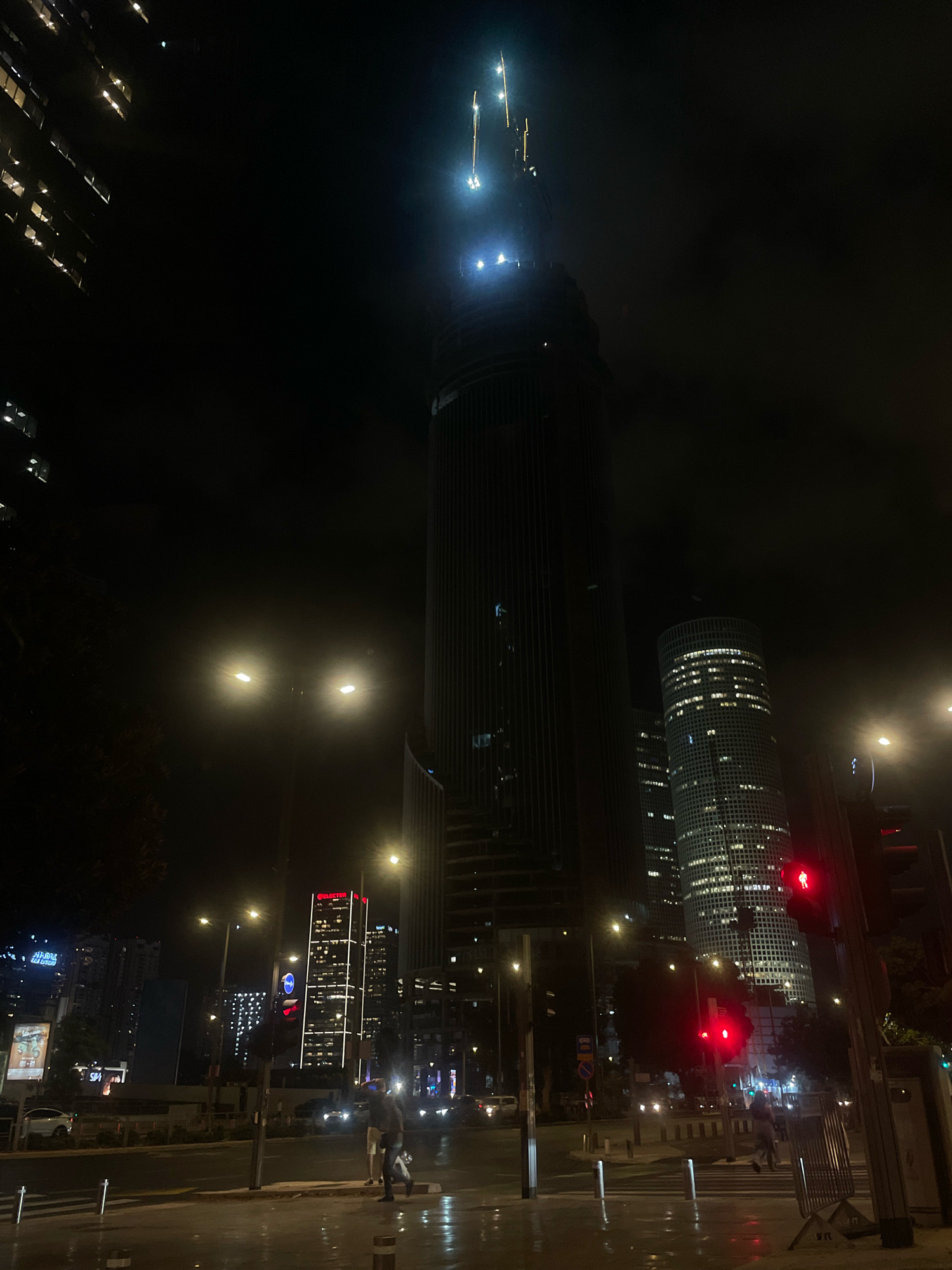 The image captures a nighttime urban scene in Tel Aviv, Israel, likely in the vicinity of the Azrieli Center. Dominating the frame is a very tall skyscraper under construction, with bright lights illuminating its upper levels and crane. To its right, the distinct circular Azrieli Center towers are visible, their windows lit, indicating occupancy. In the foreground, a wet street reflects the city lights, suggesting recent rain. Traffic lights are visible, one displaying a red signal with a pedestrian symbol, indicating a stop. Several cars are present on the street, their headlights and taillights creating streaks of light. Two individuals are walking on the wet sidewalk, one carrying a bag. Streetlights cast a warm glow, illuminating the street and sidewalk. The overall atmosphere is that of a modern, active city at night, with a focus on the impressive architecture and urban infrastructure.