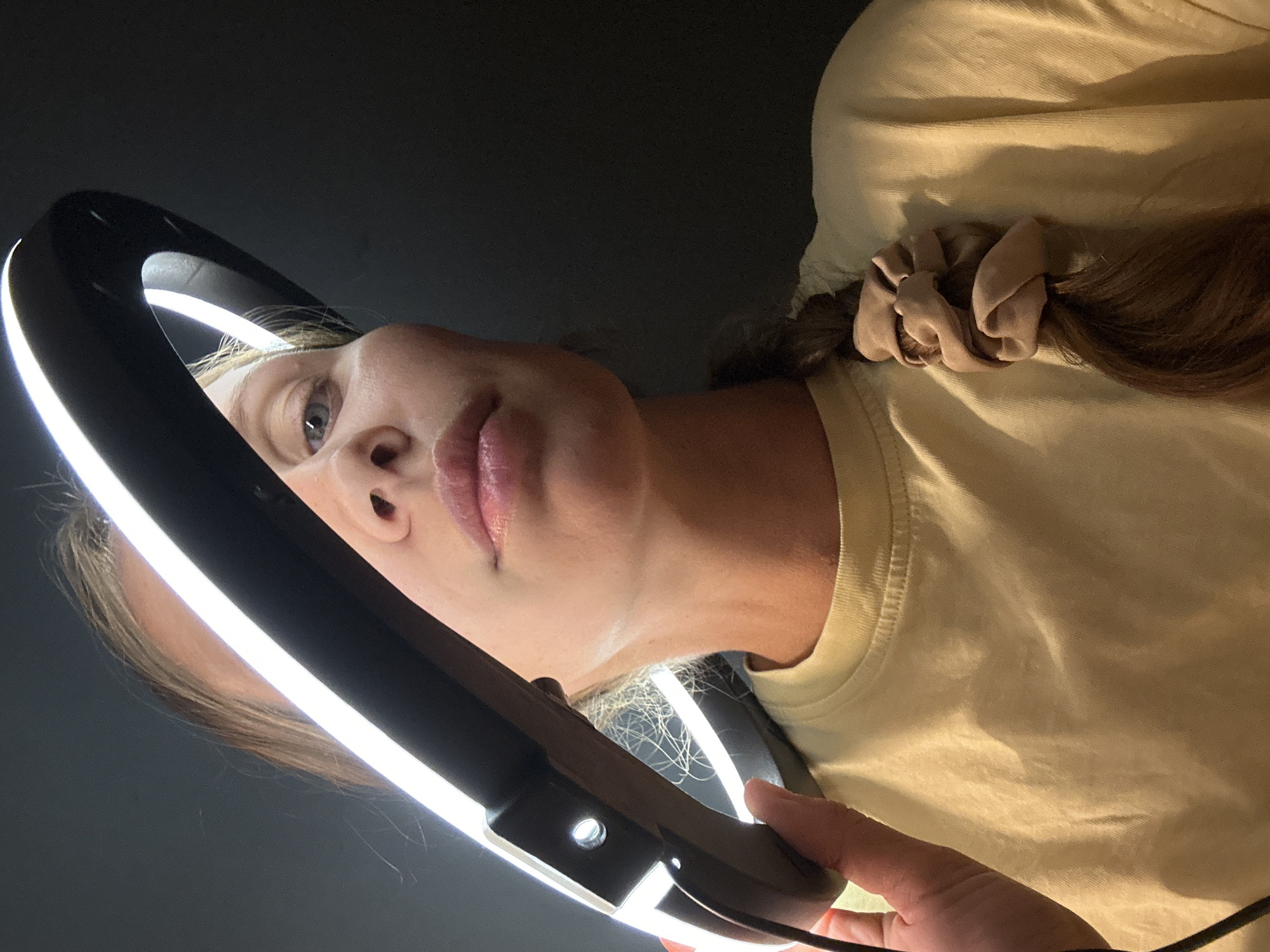 A woman is holding a ring light up to her face, apparently for self-illumination. The background is dark, possibly a room with dim lighting. The woman is wearing a light-colored shirt and has a scrunchie in her hair. The scene shows no indication of Fethiye, Turkey.