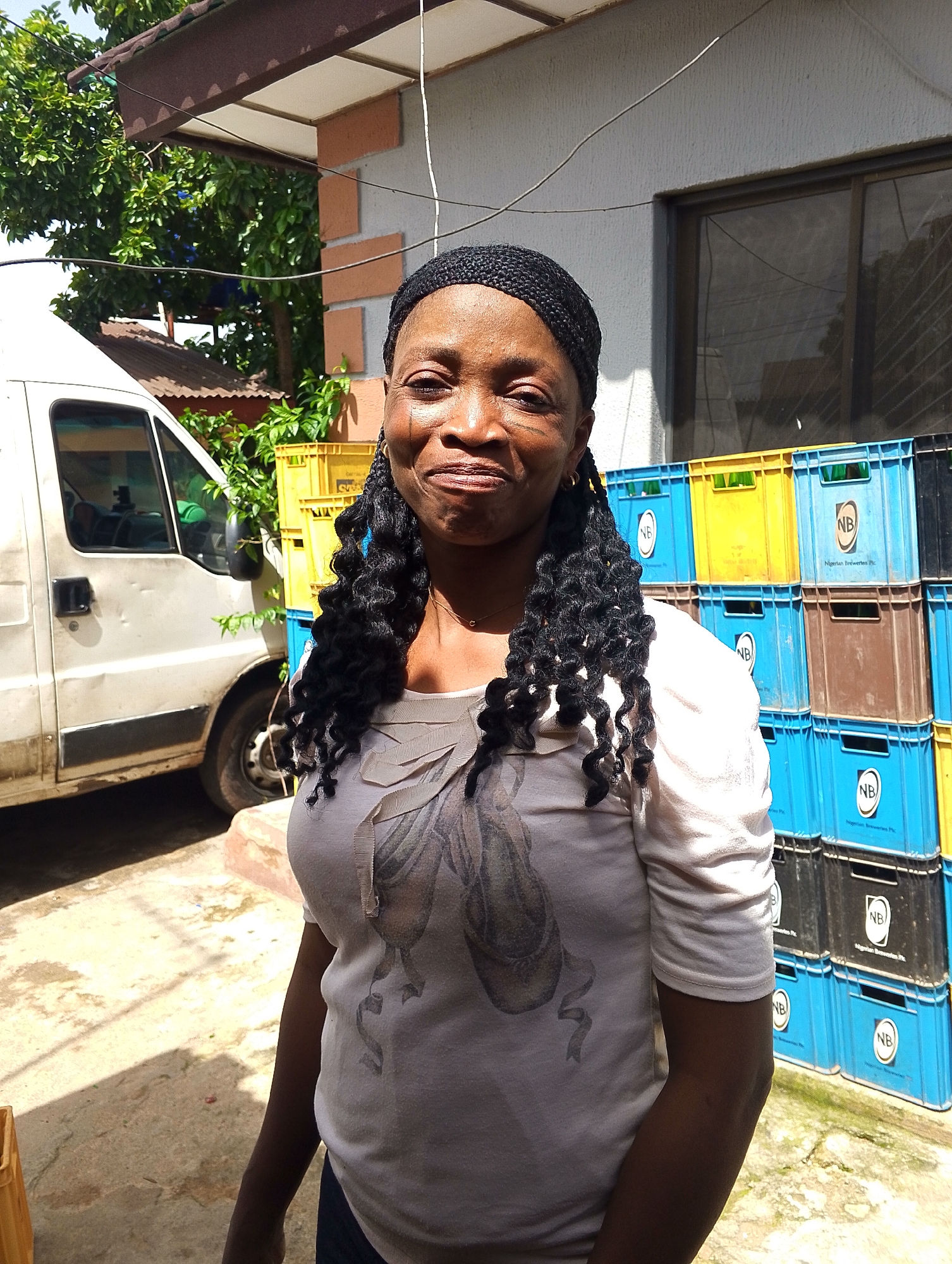 A woman with braided hair, wearing a white t-shirt with a graphic print, stands in front of a building. She has a light skin tone and is looking towards the camera with a slight smile. Behind her, to the left, is a white van. To her right and in the background, stacks of colorful crates are visible, predominantly blue and yellow, with some brown and black crates interspersed. Some of the blue crates have the "NB" logo and "Nigerian Breweries Plc." text printed on them. Green foliage is visible above and behind the crates. The ground appears to be paved with concrete, with shadows cast by the setting sun. The overall scene suggests an outdoor environment, possibly a commercial area or storage yard. The lighting indicates it is daytime, with bright sunlight. The expression on the woman's face conveys a relaxed and friendly demeanor. The setting appears to be in Jos, Nigeria, based on the provided location context.