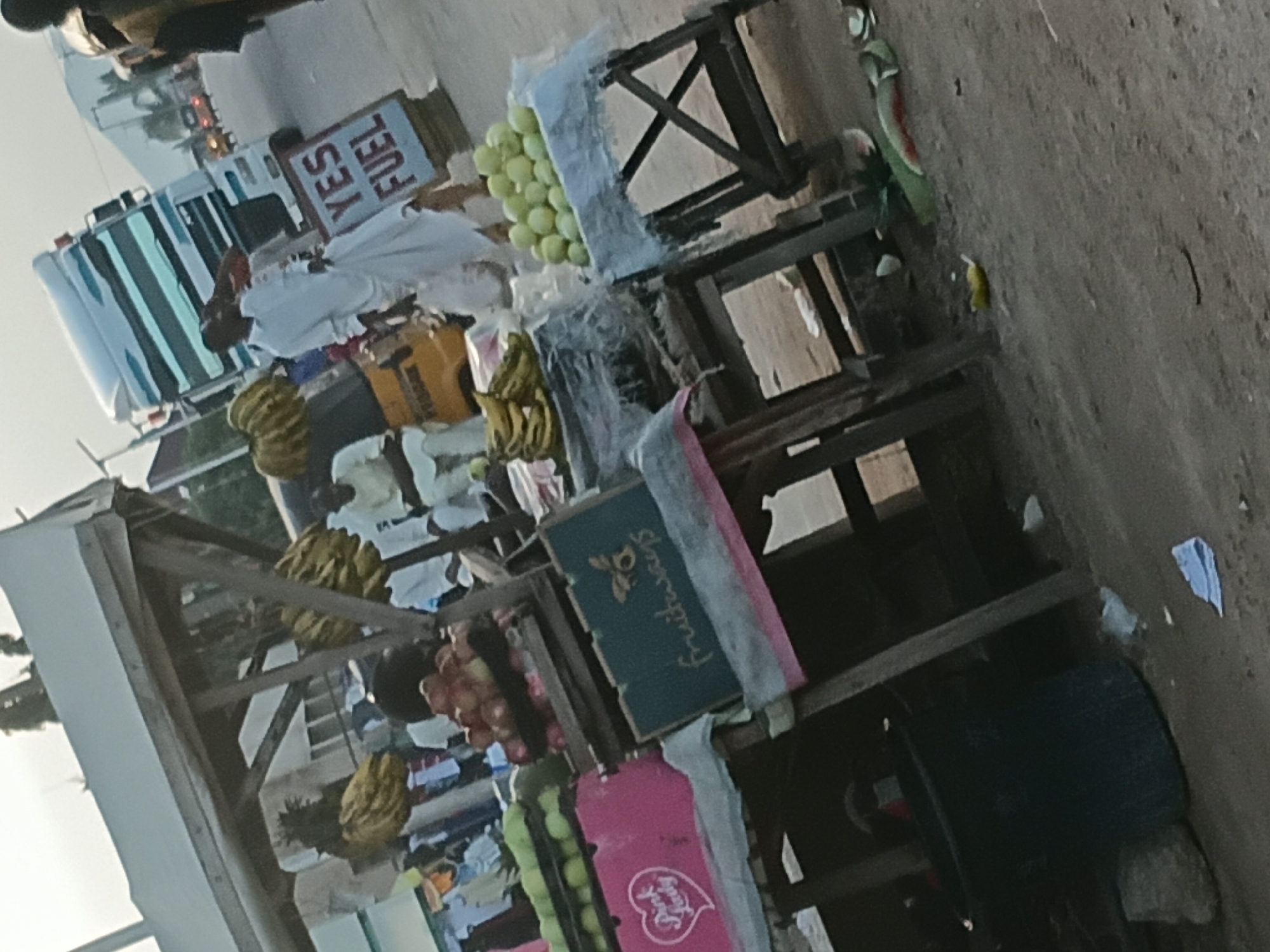 This image depicts an outdoor market scene, likely on a street, in Jalingo, Nigeria. In the foreground, there are several market stalls laden with fruits. One stall displays a pile of green fruits, possibly gooseberries, arranged on a table covered with a white cloth. Adjacent to it, a crate with the logo "fruitways" and a bee illustration is visible. Another stall has a pink box labeled "Pink Lady" with a heart graphic, containing what appears to be apples. Bunches of yellow bananas hang from a wooden structure that also supports a white canopy. In the middle ground, a man in a white robe is walking past a large white truck with blue and silver stripes. A sign nearby reads "YES FUEL". A yellow tricycle is also present. In the background, other market activities and vehicles are faintly visible, suggesting a bustling street environment. The ground is dirt and gravel, with some scattered debris. The lighting indicates it is daytime, possibly late afternoon due to the soft, diffused light.