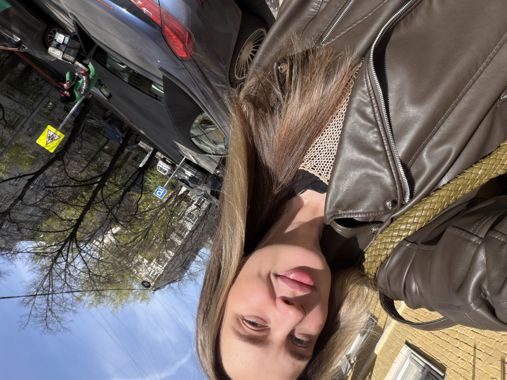The image is a selfie, rotated 90 degrees counter-clockwise, featuring a woman outdoors on a sunny day. The woman has long, light brown hair, a subtle smile, and a nose piercing. She is wearing a brown leather jacket over a black top with a visible mesh texture around the neckline. A green braided strap, likely from a bag, is visible over her right shoulder.
In the background, a dark blue or grey sedan is parked, displaying a Russian license plate with the alphanumeric combination "B71043" and the region code "199", indicating Moscow. Adjacent to the car, a yellow triangular road sign shows a black image of children and the Cyrillic word "школа" (school) beneath it, signifying a school zone. Further in the background, a blue rectangular sign with a white "P" denotes a parking area.
The scene depicts a city street during what appears to be spring, with trees beginning to bud green leaves. Buildings with light-colored facades are visible against a bright blue sky with a few clouds. The strong sunlight creates clear shadows, suggesting it is midday or afternoon. A yellow brick building is visible in the lower right portion of the background.