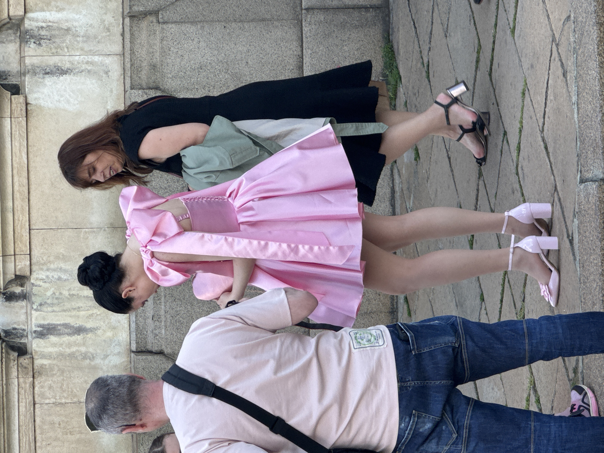 The image depicts three people on a paved surface, possibly a street or plaza. In the foreground, a man wearing a pale pink shirt, dark blue jeans, and a black shoulder strap is partially visible. To his right is a pink and black running shoe. Above him, a woman in a pink satin dress stands next to a woman in a black dress. The woman in pink is wearing pink block-heeled shoes with ankle straps. The woman in black is wearing black heeled sandals. A light green jacket is slung over her left shoulder. The background features a stone wall. The location is likely in Sofia, Bulgaria.
