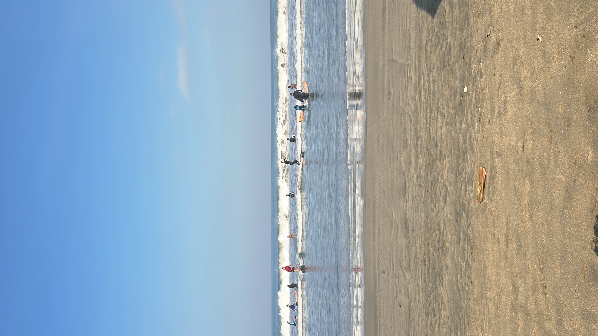 The image shows a beach scene in Kuta, Indonesia. The sky is clear blue. The ocean has small waves with people surfing. The beach is sandy and light brown, with one sandal lying on the sand.