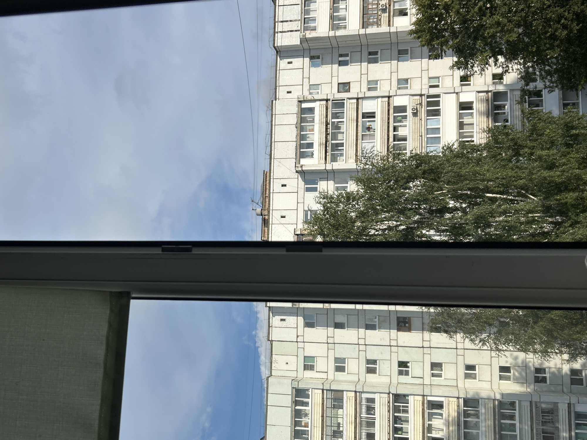 The image depicts a view from a window overlooking a residential building in Chaikovsky, Russia. The building is a multi-story apartment block, characterized by its light-colored facade, numerous windows, and balconies. Trees, some with white bark, are visible in front of the building, partially obscuring the lower floors. The sky above is partly cloudy, with patches of blue visible. A window frame and a roller blind are present in the foreground, suggesting the perspective is from inside a room. There are no people or specific actions clearly identifiable in the scene. The overall impression is that of a typical urban residential environment on a daytime.