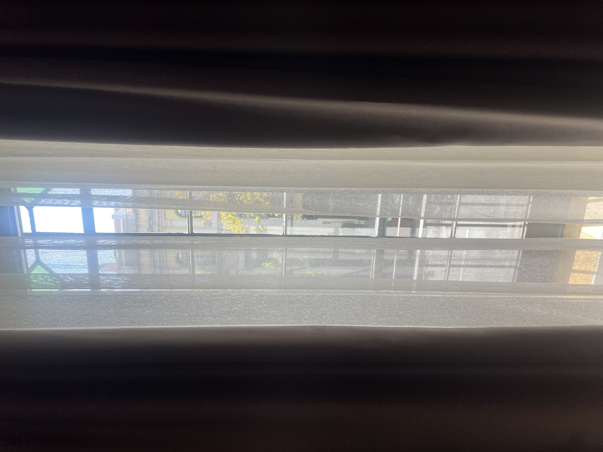 This is a medium shot of a window in Sevilla, Spain. The window is framed by dark brown curtains on either side and a sheer white curtain in the center. Through the window, a building with an arched doorway and yellow facade is visible, along with a tree with yellow leaves. The window itself has a metal grate, indicating it is on an upper floor. The light suggests it is daytime.