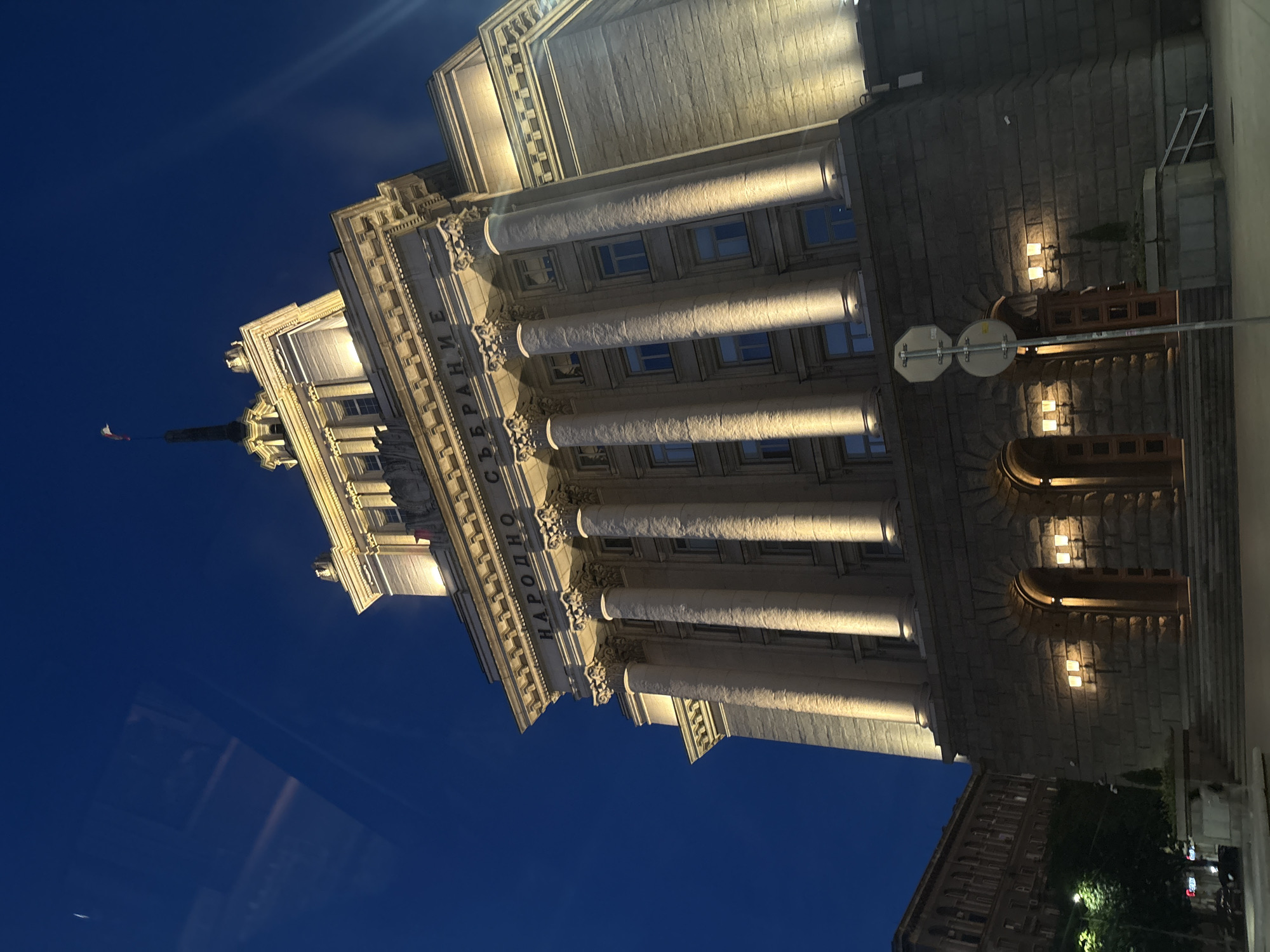 A low-angle, wide shot captures the facade of a grand, neoclassical building at dusk. The building is constructed of stone with large, illuminated columns and intricate architectural details. The sky above is a deep, dark blue, suggesting twilight or night. The building's windows are dark, but several exterior lights are illuminated, casting a warm glow on the facade. In the distance, on the left side of the image, another building with similar architectural style is visible, along with some trees and streetlights. The overall scene conveys a sense of historical grandeur and stillness.