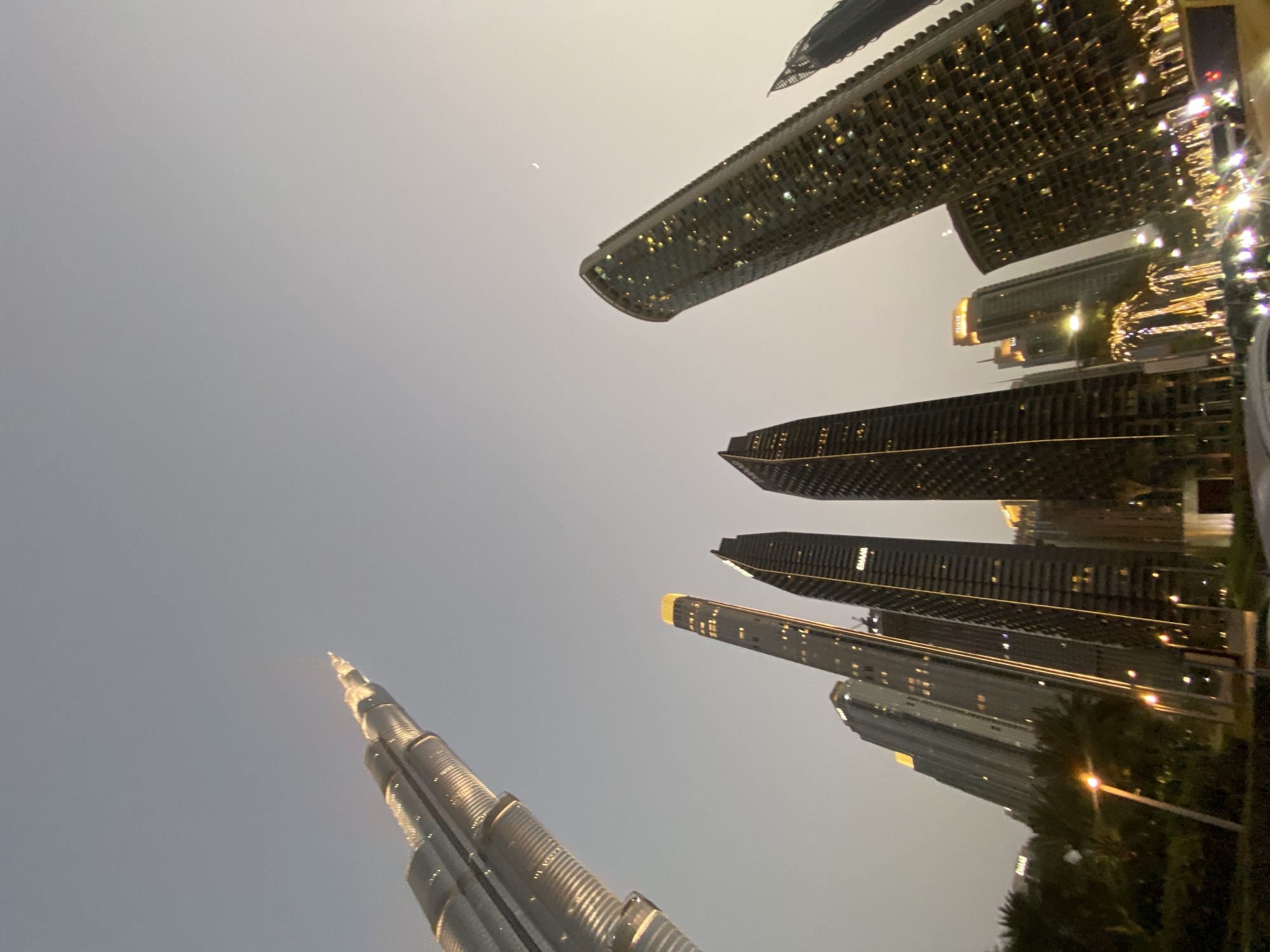 The image captures a low-angle view of the Dubai skyline at dusk. The Burj Khalifa is prominently displayed on the left, stretching upwards. Several other skyscrapers are visible, some illuminated with interior lights. The sky is a pale grey, and there is a crescent moon. Some foliage and street lighting can be seen at the bottom of the frame.