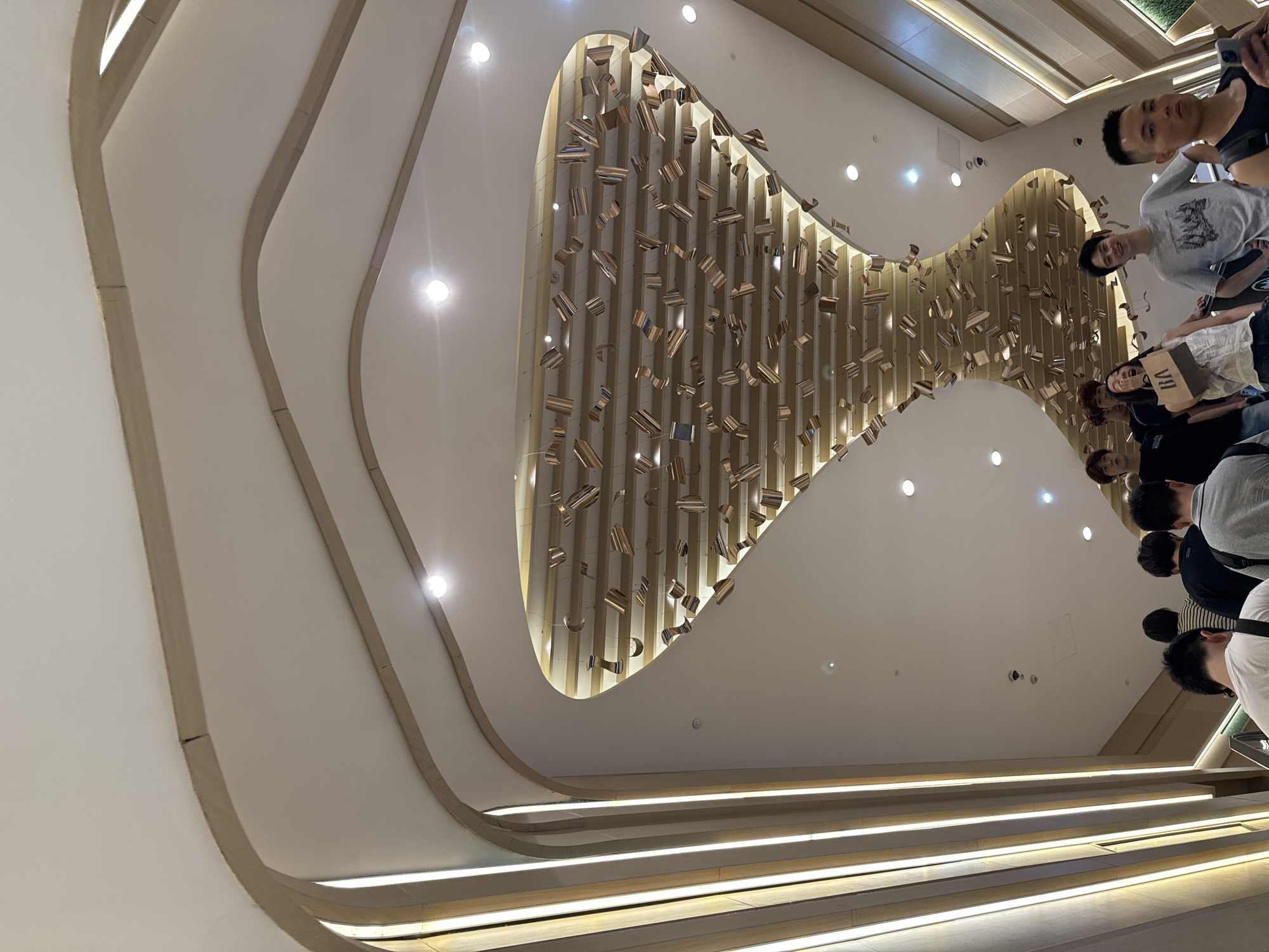 The image depicts a group of young adults on an escalator ascending within a brightly lit, modern interior space. The architecture features a curved, beige-toned ceiling with integrated vertical lighting strips along the left side. A prominent decorative element is suspended from the ceiling above the escalator; it consists of numerous small, reflective rectangular objects arranged in a seemingly random pattern, creating a visually striking effect. Several individuals are visible on the escalator, appearing to be in casual conversation or looking around. The overall ambiance suggests a shopping mall or a similar public commercial area in a city like Liangjiang, China, characterized by contemporary design and a vibrant atmosphere.