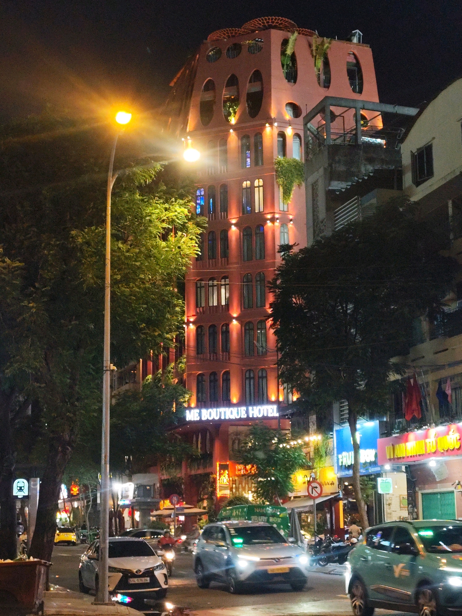 This nighttime street scene captures the facade of the "ME BOUTIQUE HOTEL" in Ho Chi Minh City, Vietnam. The hotel building, painted a distinctive coral pink, features a unique architectural design with oval-shaped openings and balconies adorned with hanging plants. The building is illuminated by orange lights. A street lamp with a bright yellow light dominates the left side of the frame, casting a warm glow. Several cars are visible on the street, including a silver sedan with its headlights on, a light blue electric car, and a yellow taxi in the distance. The street is lined with trees and other buildings, some of which are also illuminated. Signs in Vietnamese are visible on some of the storefronts. The scene depicts a bustling urban environment at night.